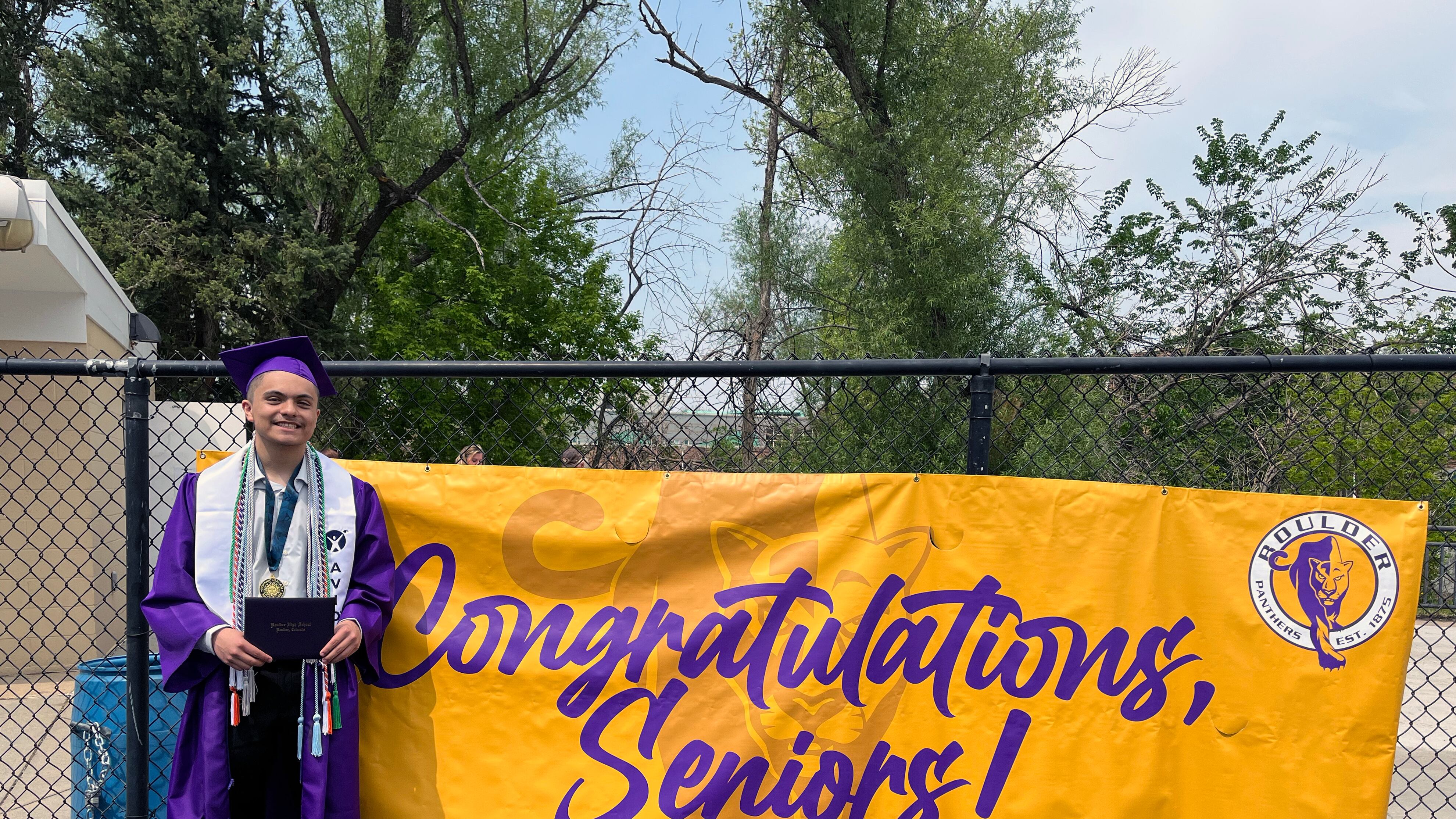 A high school senior wears dark purple graduation gown and hat while holding a diploma next to a large yellow sign that reads "Congratulations, Seniors!"
