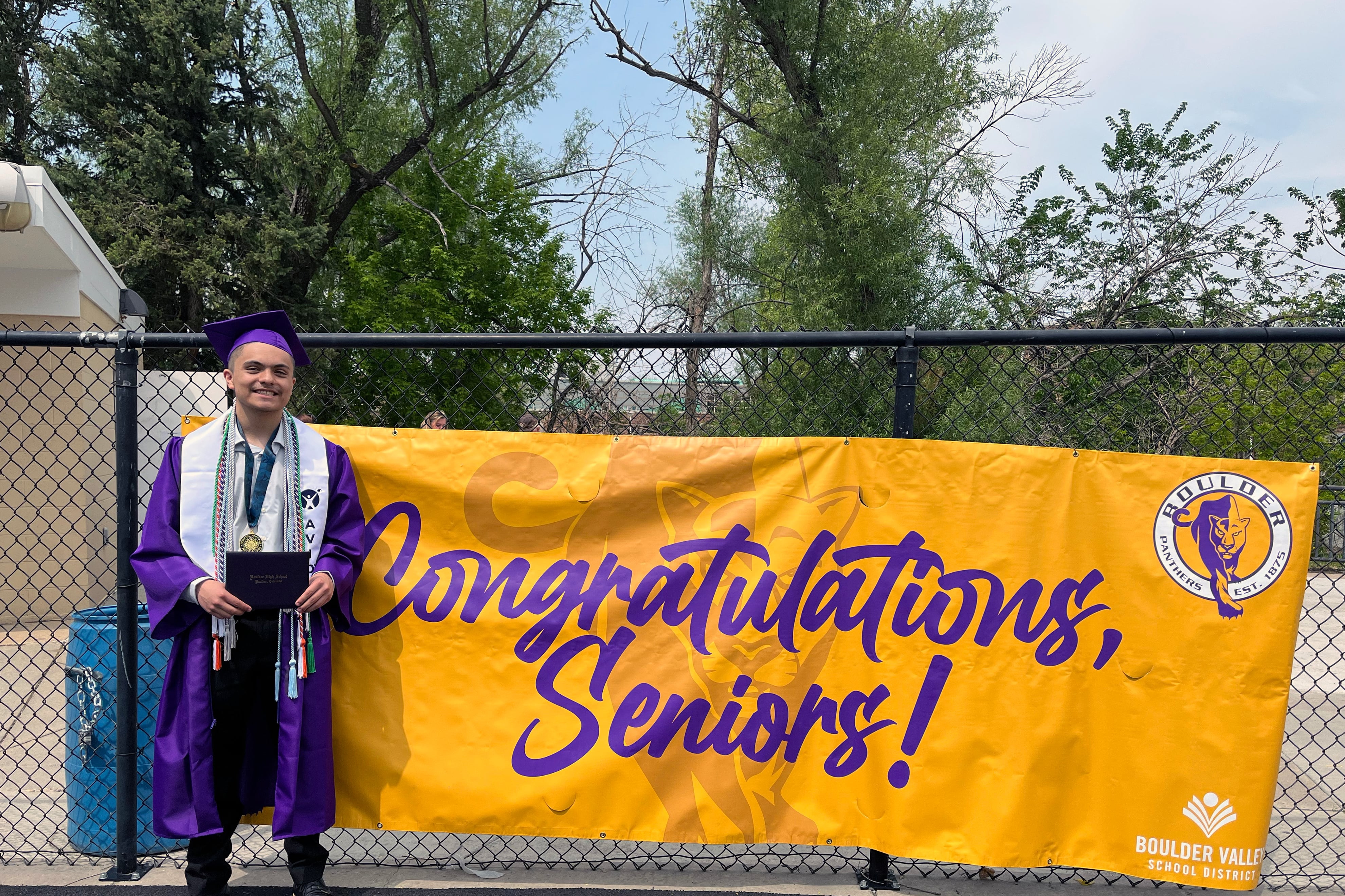 A high school senior wears dark purple graduation gown and hat while holding a diploma next to a large yellow sign that reads "Congratulations, Seniors!"