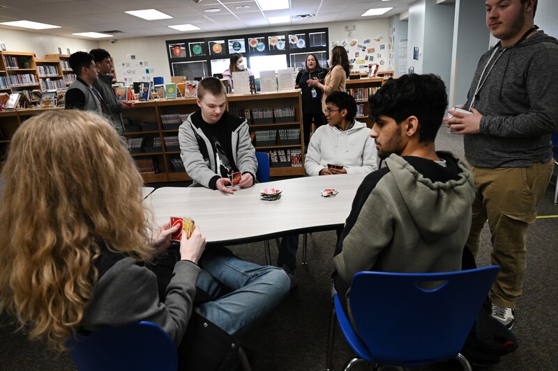 A group of high school students play a card game at a table with bookshelves in the background.