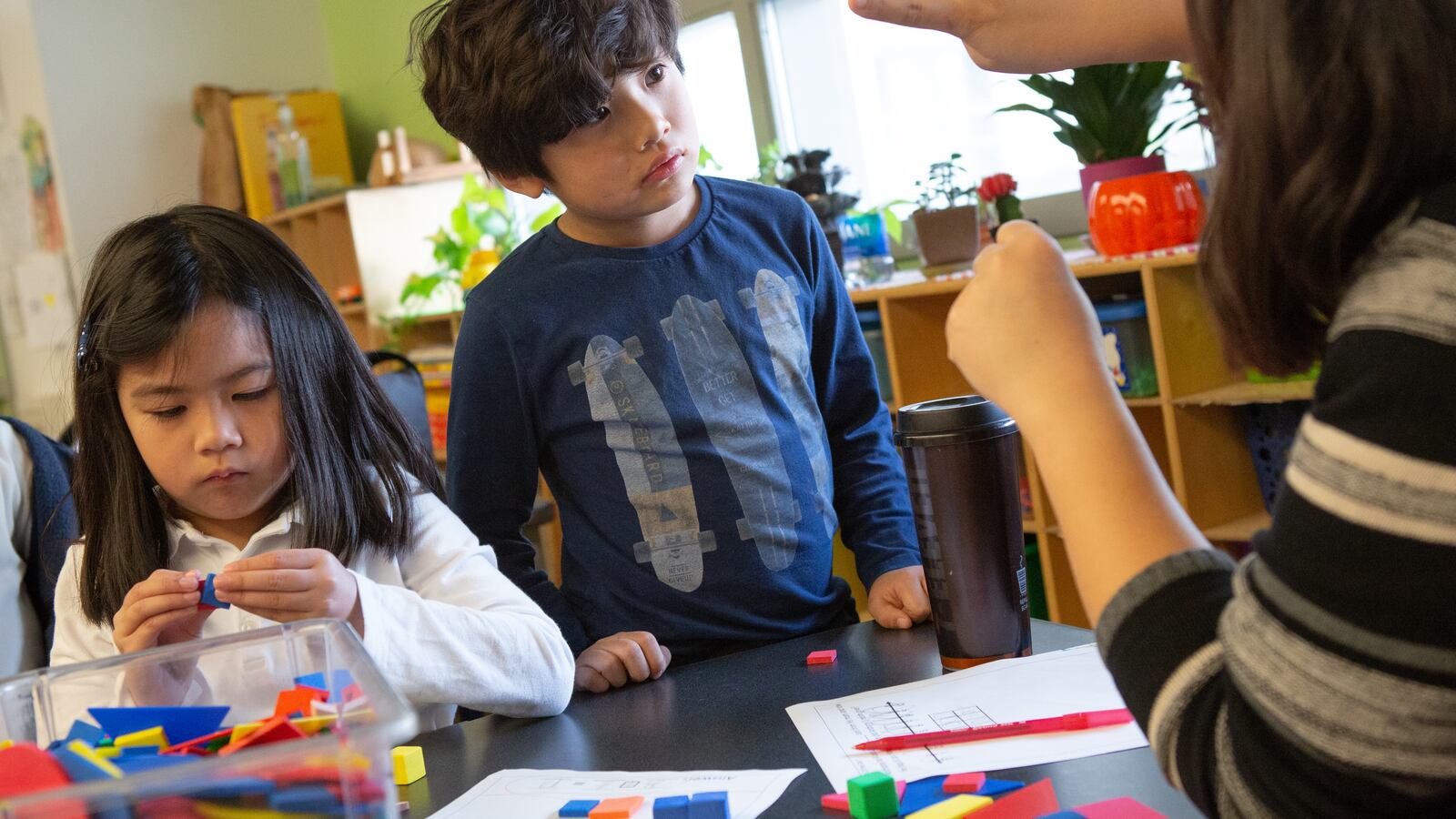 A teacher in the far right of the image holds up four fingers for a young boy who concentrates on counting them. To the left, a girl works on a project at the same table.