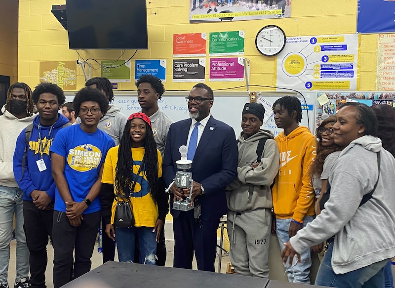 A man in a suit stands in the middle of a group of high school students holding a robot while posing for a photo.