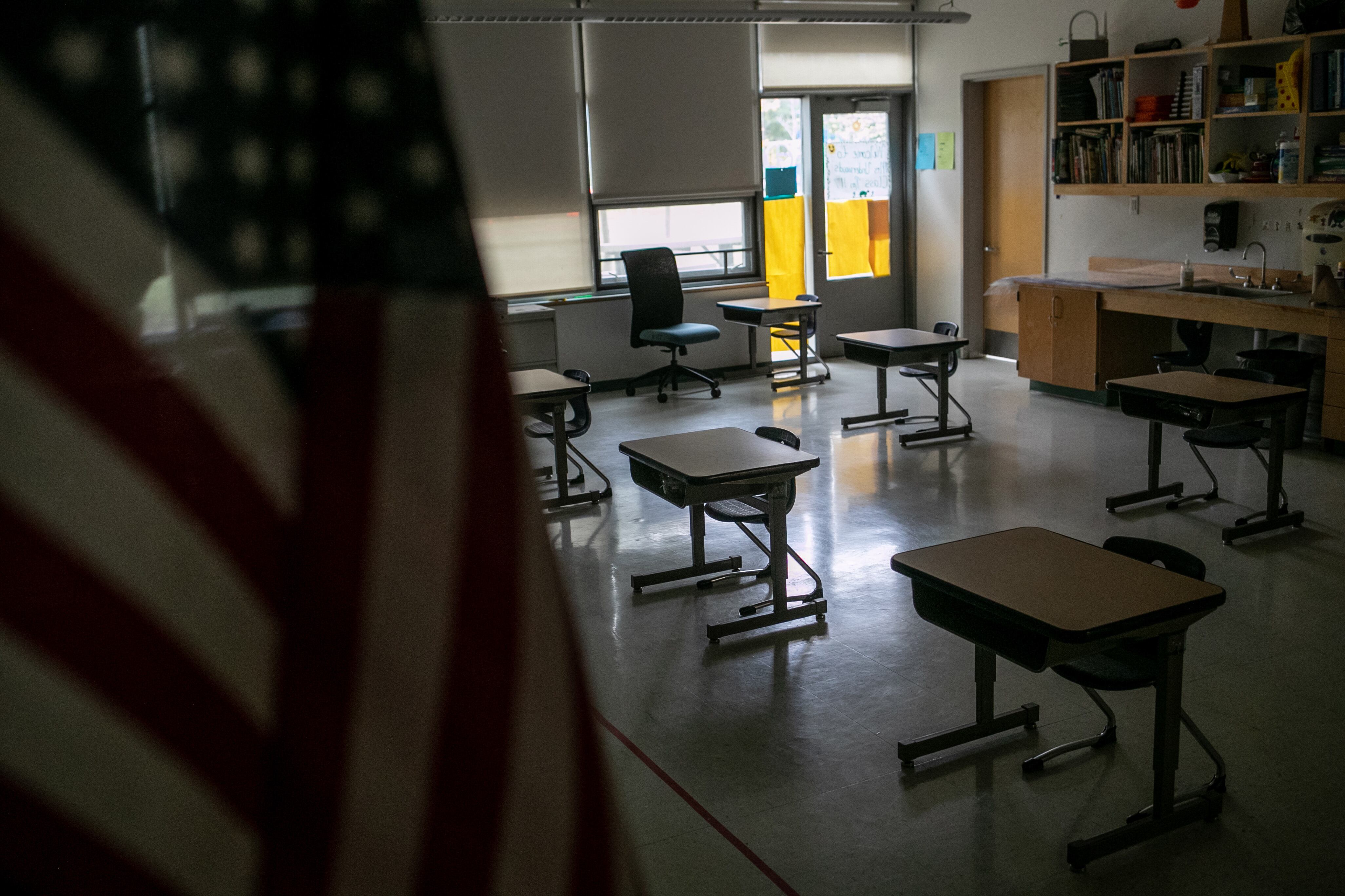 A photo with an American flag in front with a row of desks in a dimly lit classroom.