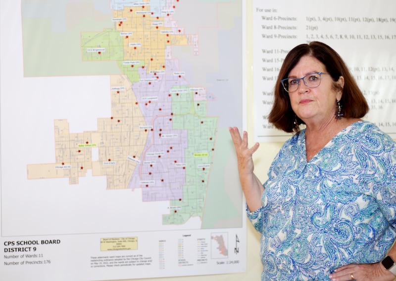 A woman wearing a blue blouse stands in front of a pastel colored map hanging on a wall.