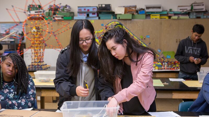 Two young female students with long, dark hair stand at a wooden desk working on a school project. There are two students in the background.