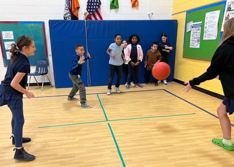 A group of students and a teacher play foursquare in a school gym.
