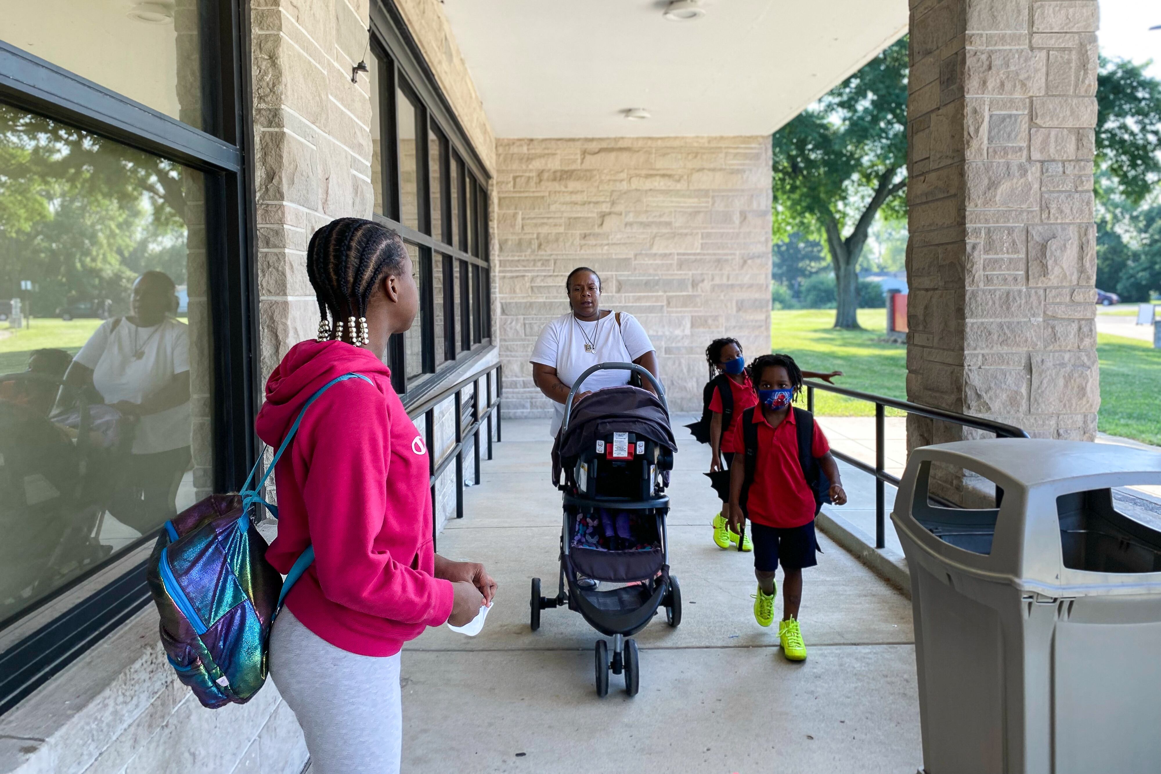 A parent walks pushing a stroller walks up a school ramp with two of her first graders and an older student wearing a backpack.