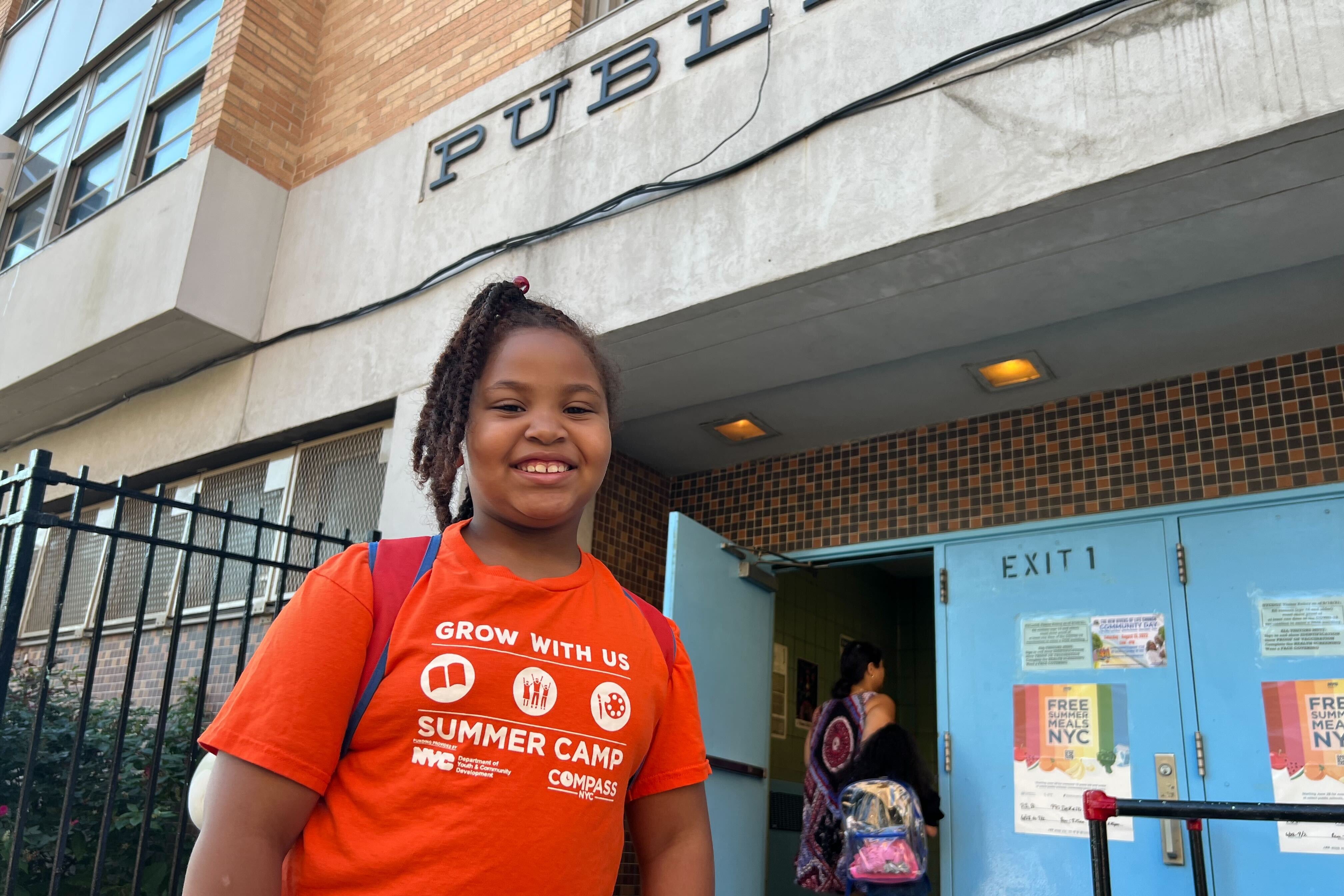 A young girl in a bright orange T-shirt stands in front of her blue school doors.