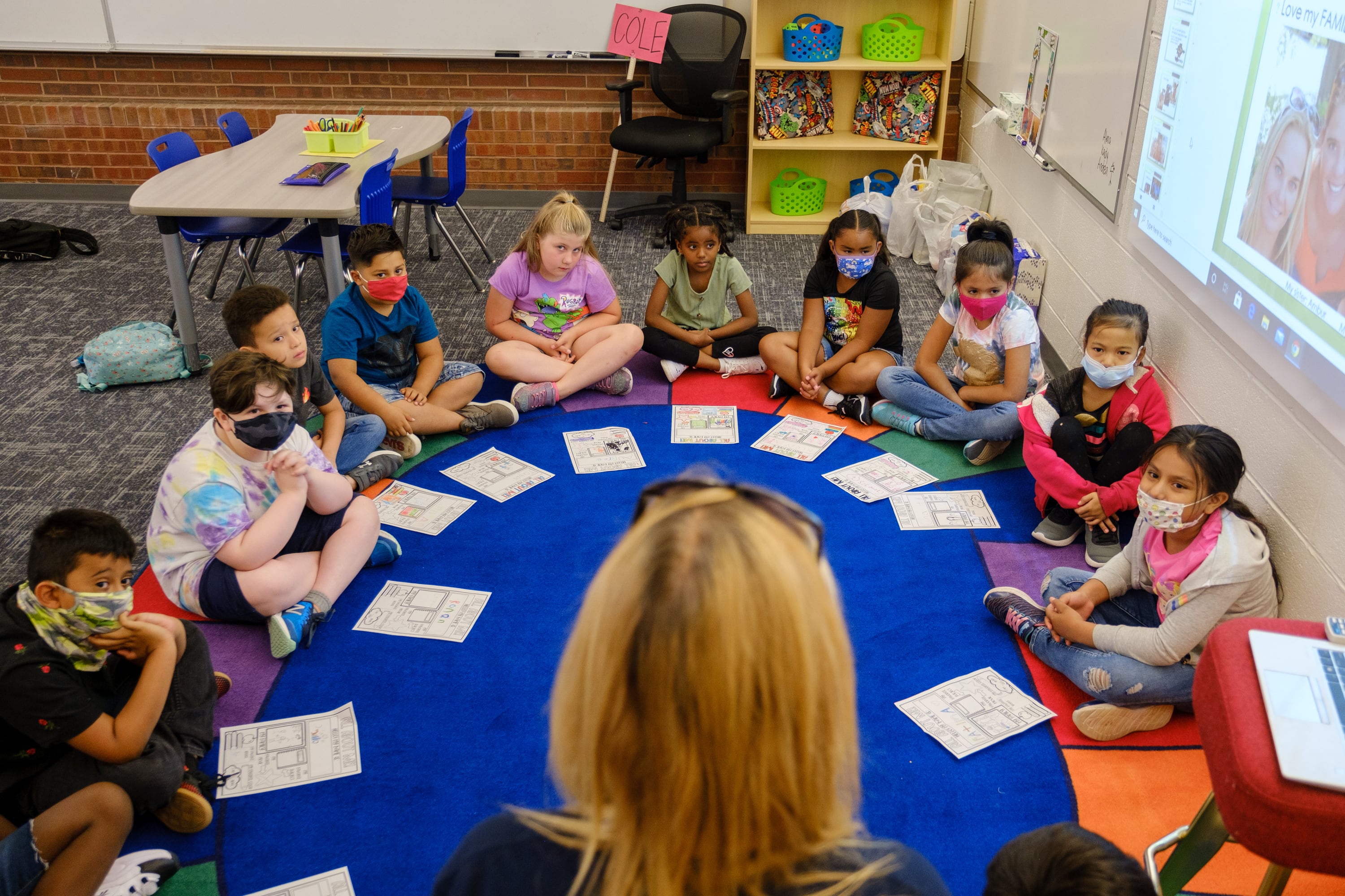 A teacher leads an introductory exercise with students sitting crossed legs in a circle on a colorful mat.