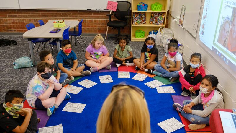 A teacher leads an introductory exercise with students sitting crossed legs in a circle on a colorful mat.