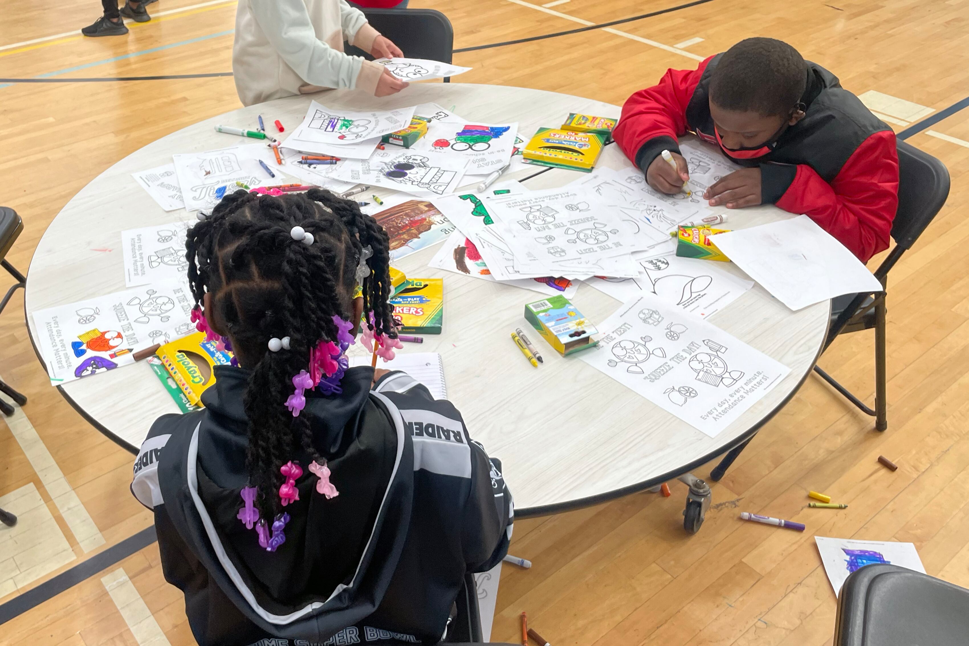 A bird’s eye view of three students seated around a round table, working with coloring pages and markers.