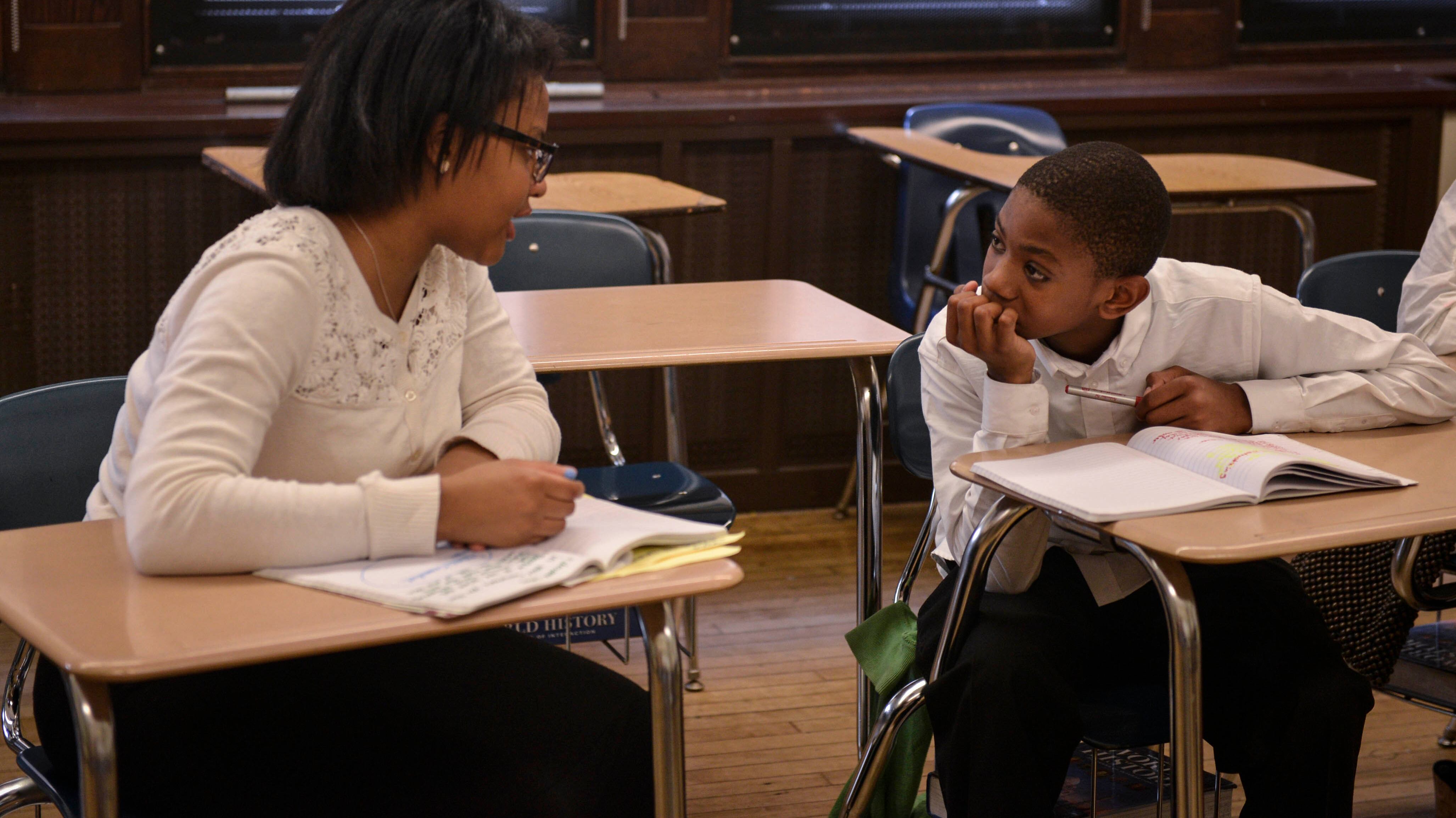 A student and a teacher sit at desks and talk to each other in a classroom while workbooks are open in front of them.