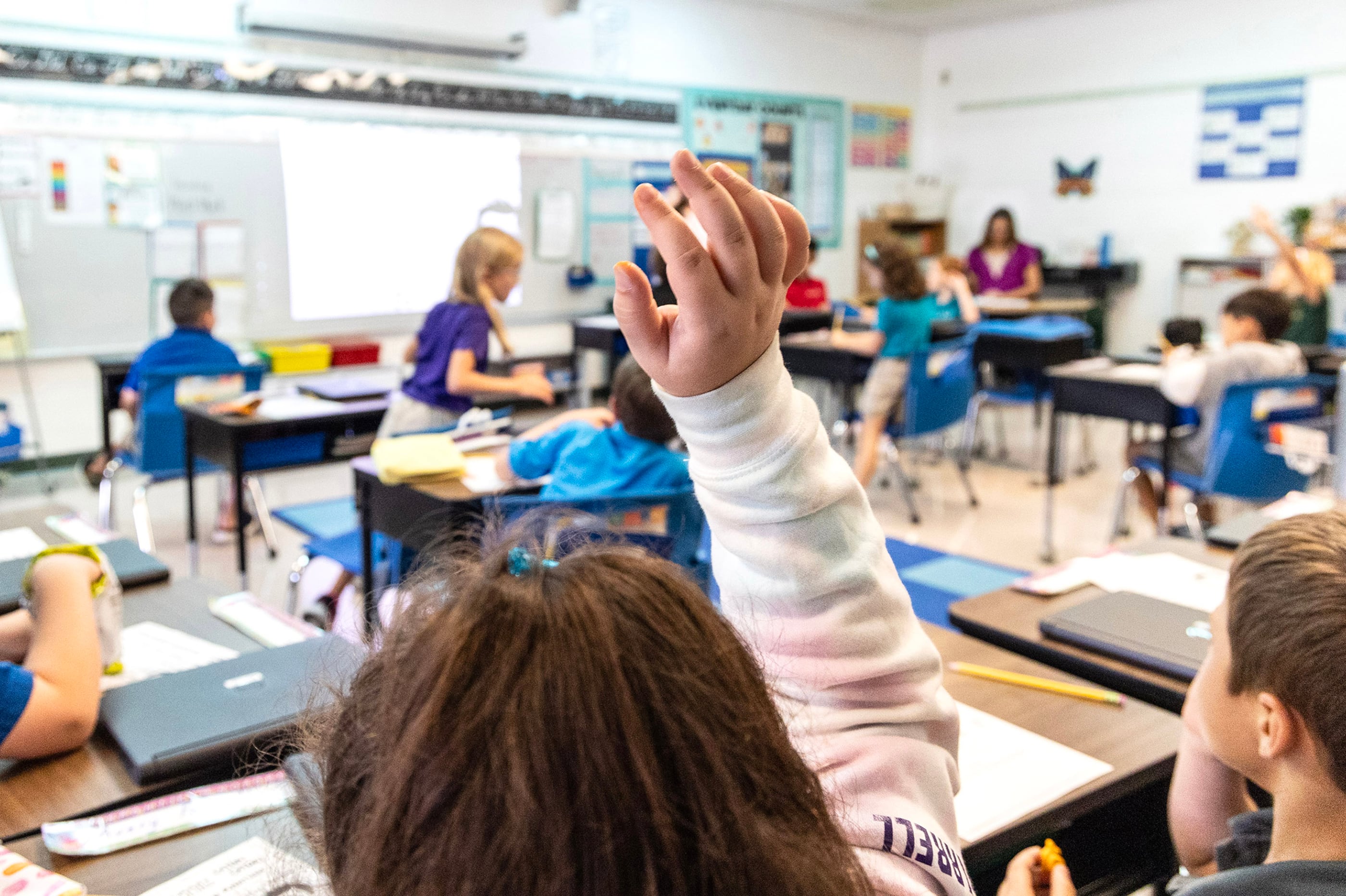 A photograph of a classroom full of students sitting at their desks with a the top of a student's head and her raised hand.