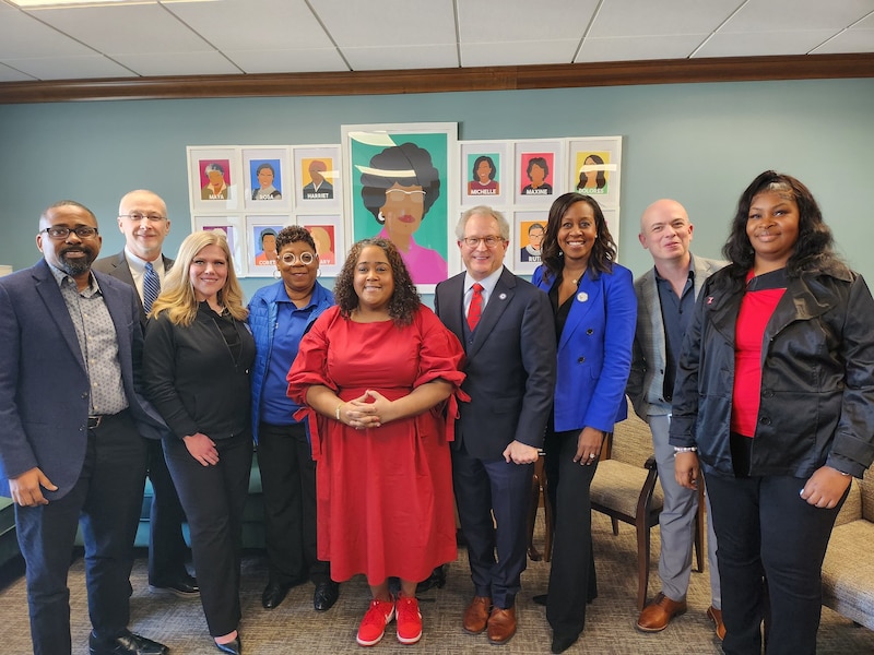 Nine people wearing business clothes stand and pose for a portrait with artwork on the wall in the background.