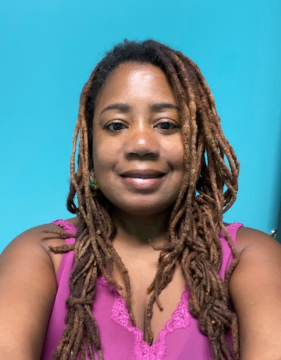 A woman wearing a pink tank top and long brown braids smiles for a portrait in front of a blue background.
