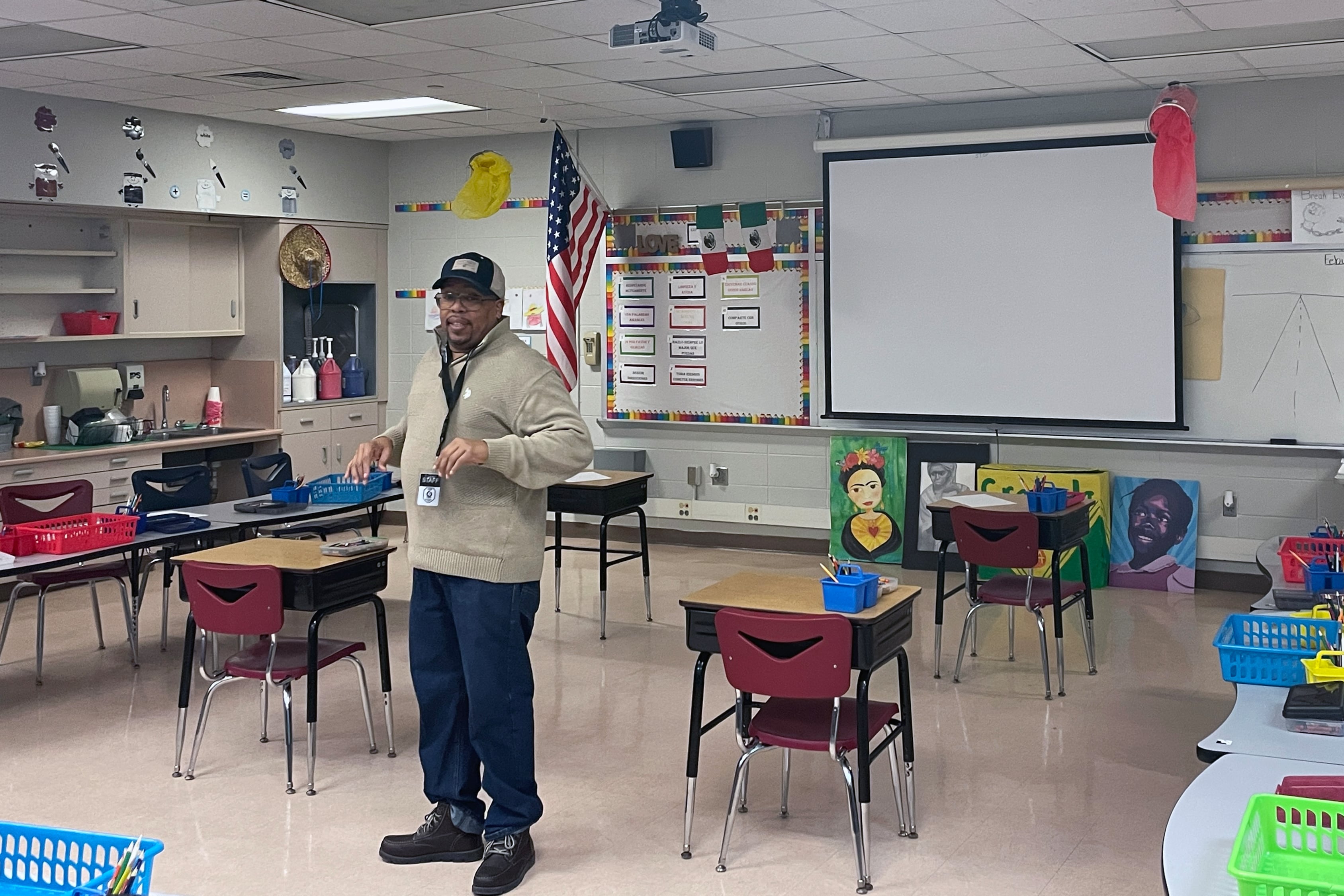 A man wearing a sweater and a baseball cap stands in front of a colorful classroom.
