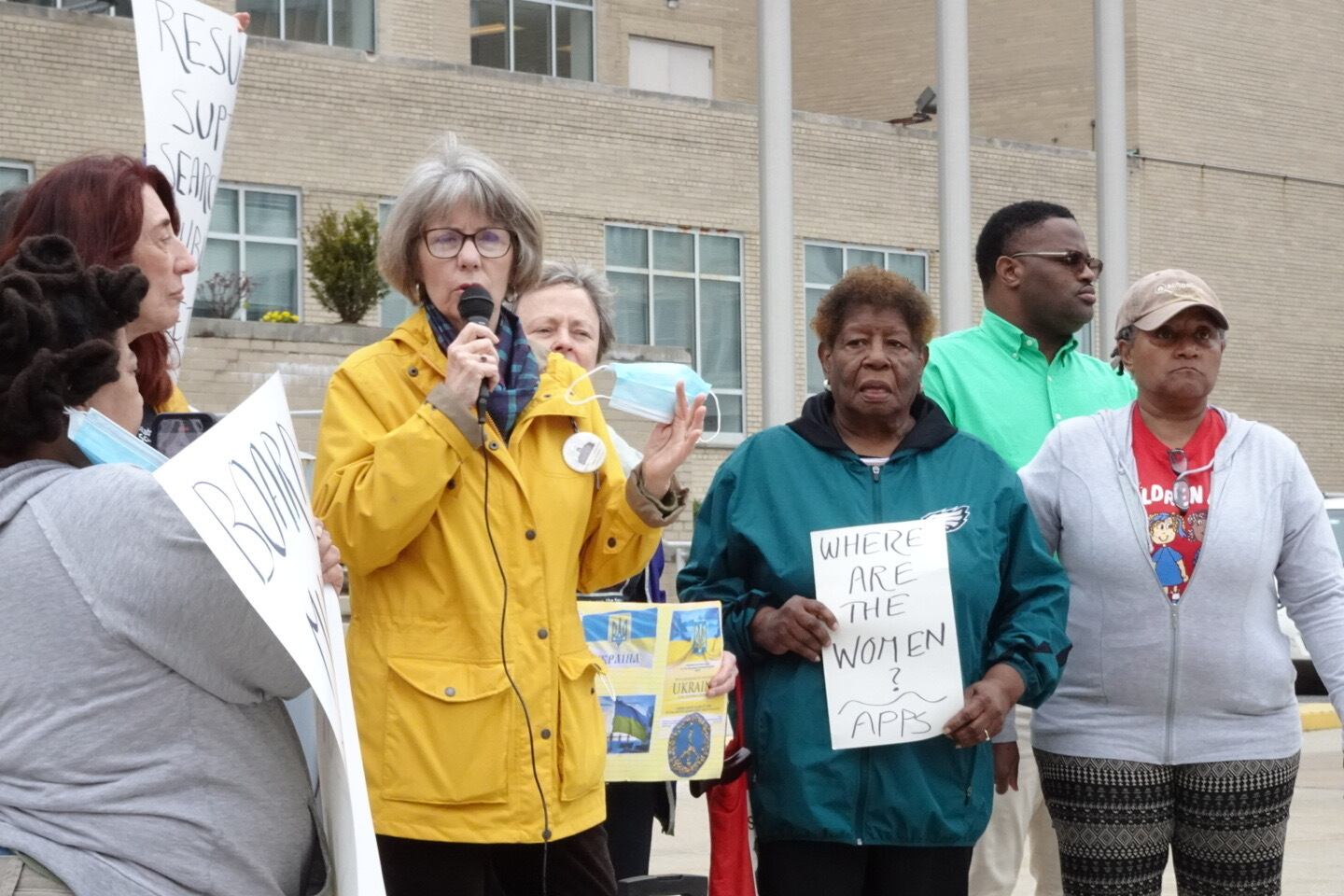 A woman in a yellow coat speaks into a microphone in front of a brick building, while several other people stand to her left and right. One woman is holding a sign that says, “Where are the women? AAPS.”