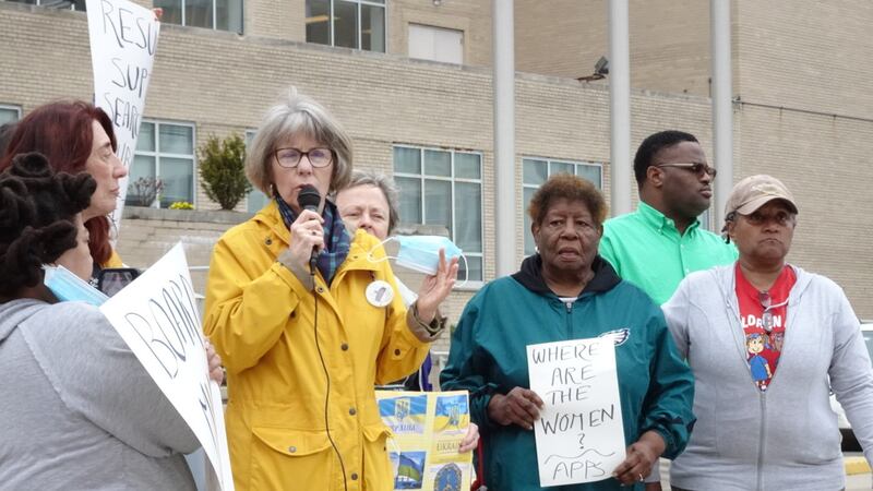 A woman in a yellow coat speaks into a microphone in front of a brick building, while several other people stand to her left and right. One woman is holding a sign that says, “Where are the women? AAPS.”
