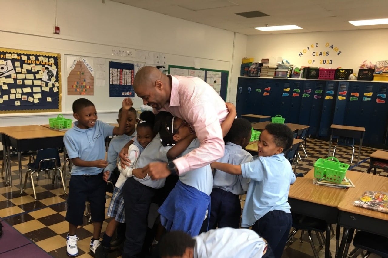 In a classroom with a checker board floor, several smiling elementary school students gather around their teacher to share a group hug.