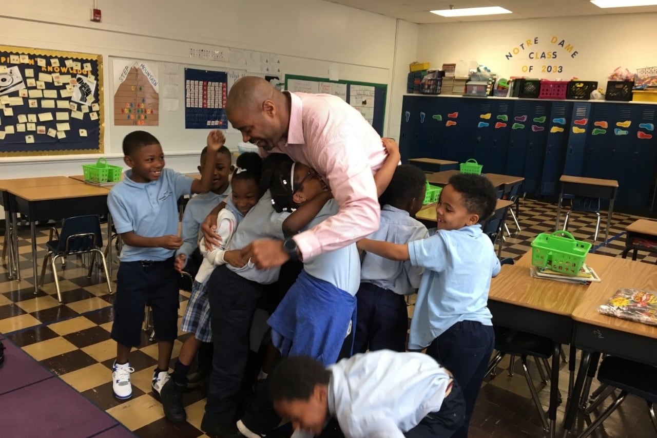 In a classroom with a checker board floor, several smiling elementary school students gather around their teacher to share a group hug.