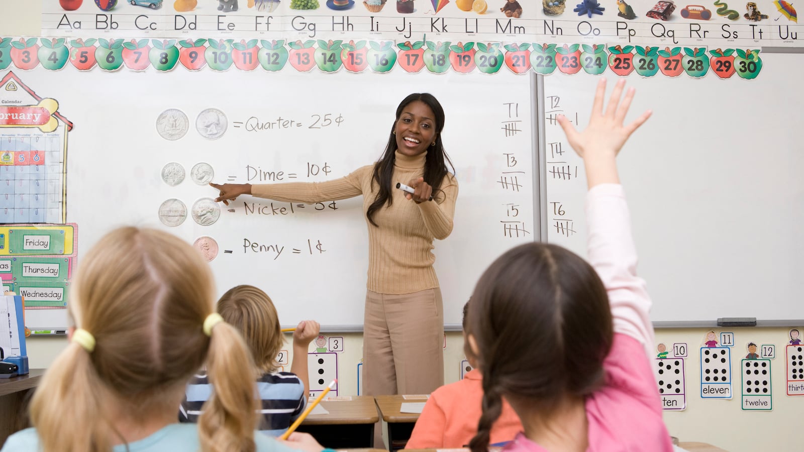 Teacher in beige outfit stands in front of a white board and points to a student in her class. 
