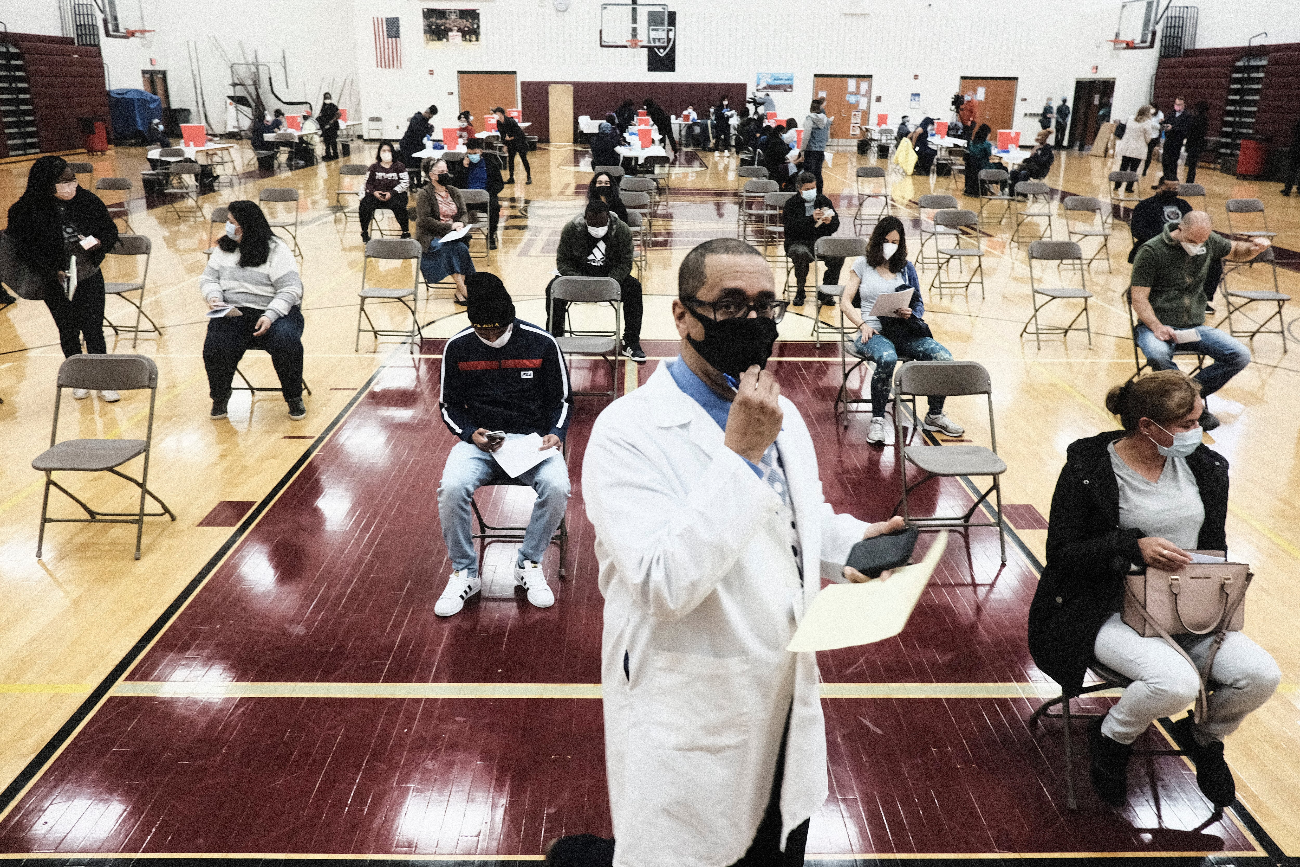 A doctor stands in front of rows of patients seated in a school gymnasium.