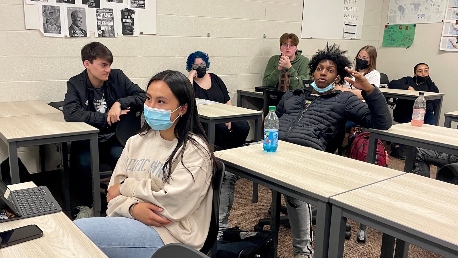Seven high school students participate in a class discussion at their desks in a classroom with white walls decorated with maps and photographs of book covers.