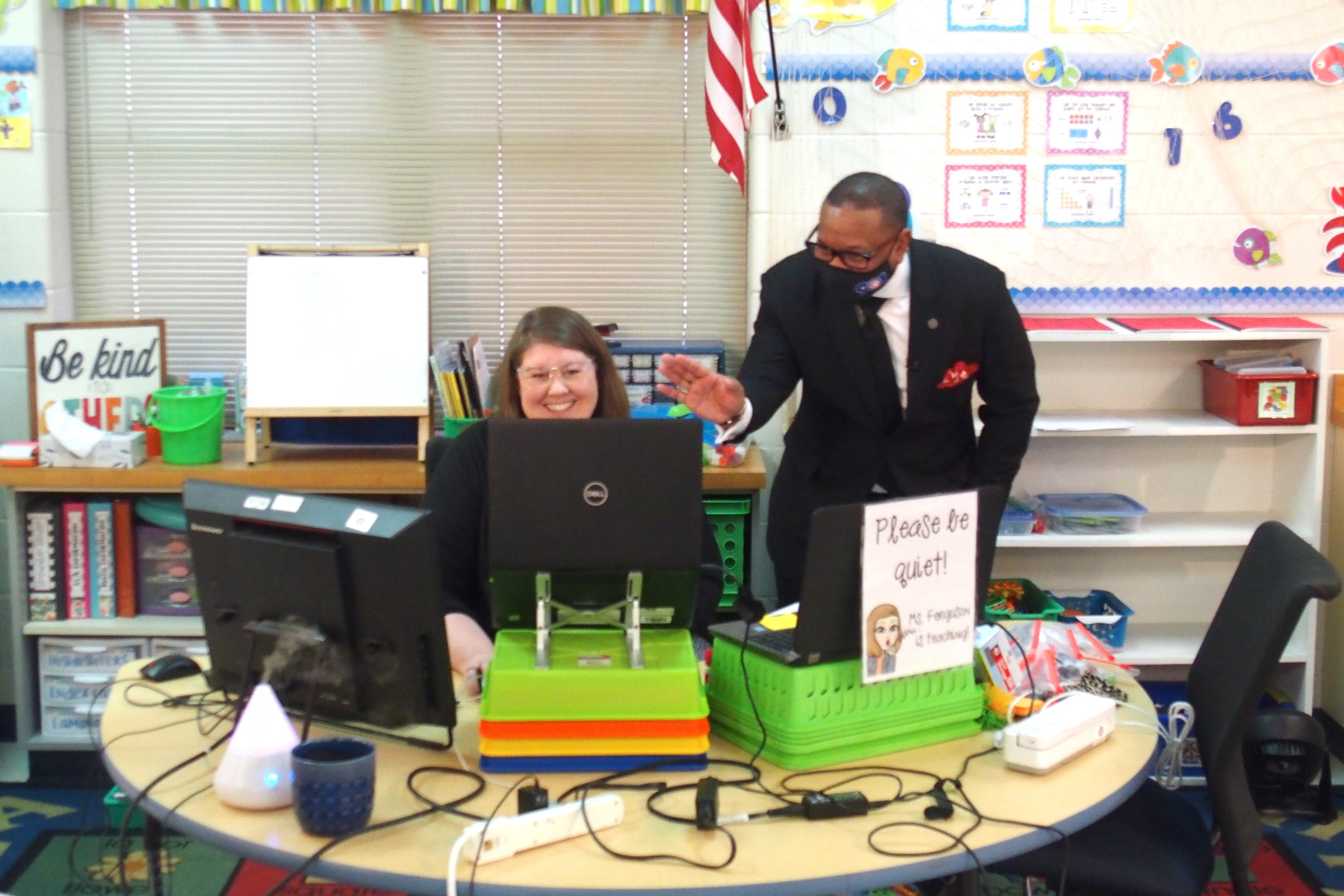 Superintendent Joris Ray bends down to wave at kindergarten students on a laptop screen who are in an online class while their teacher smiles at her desk.