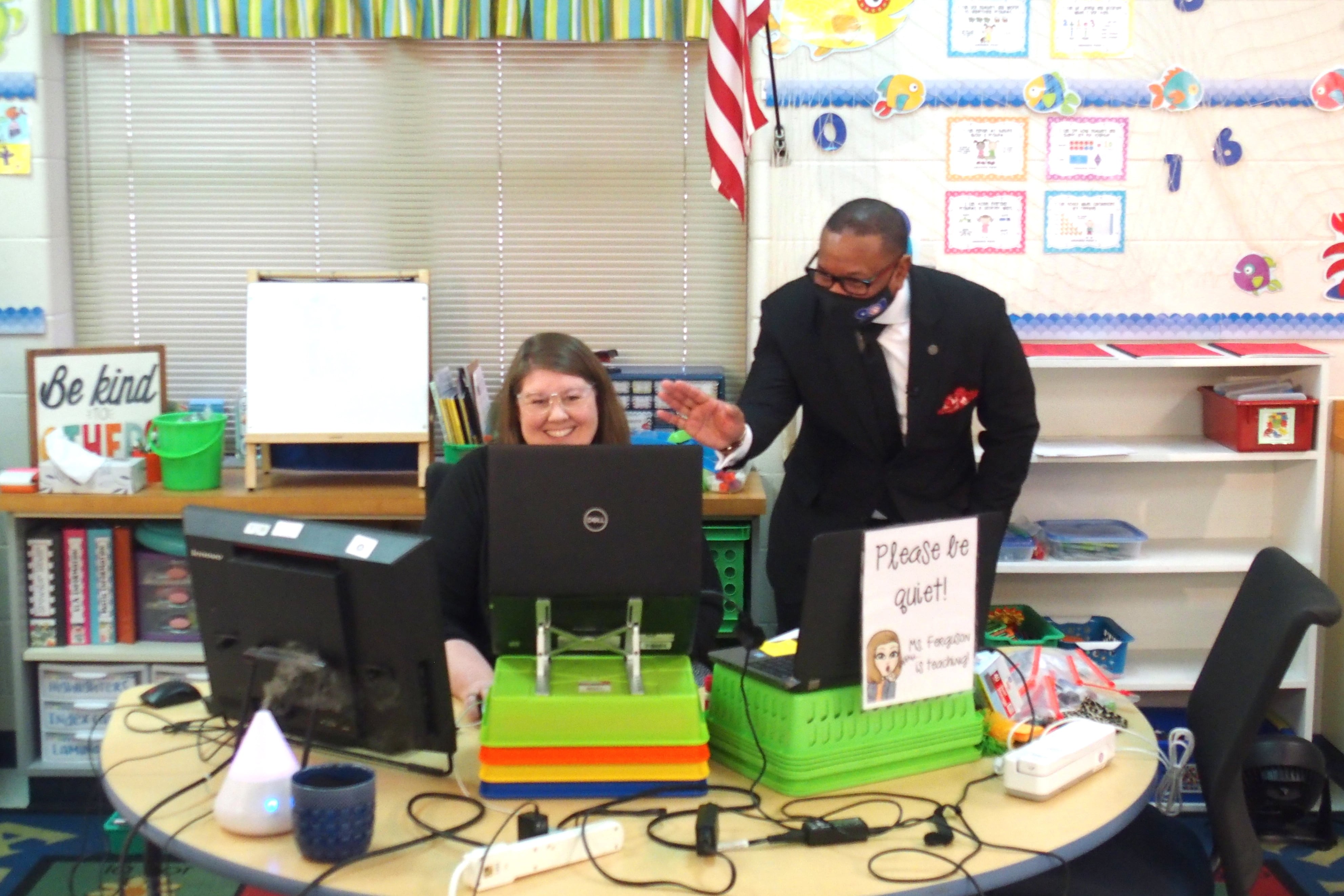 Superintendent Joris Ray bends down to wave at kindergarten students on a laptop screen who are in an online class while their teacher smiles at her desk.