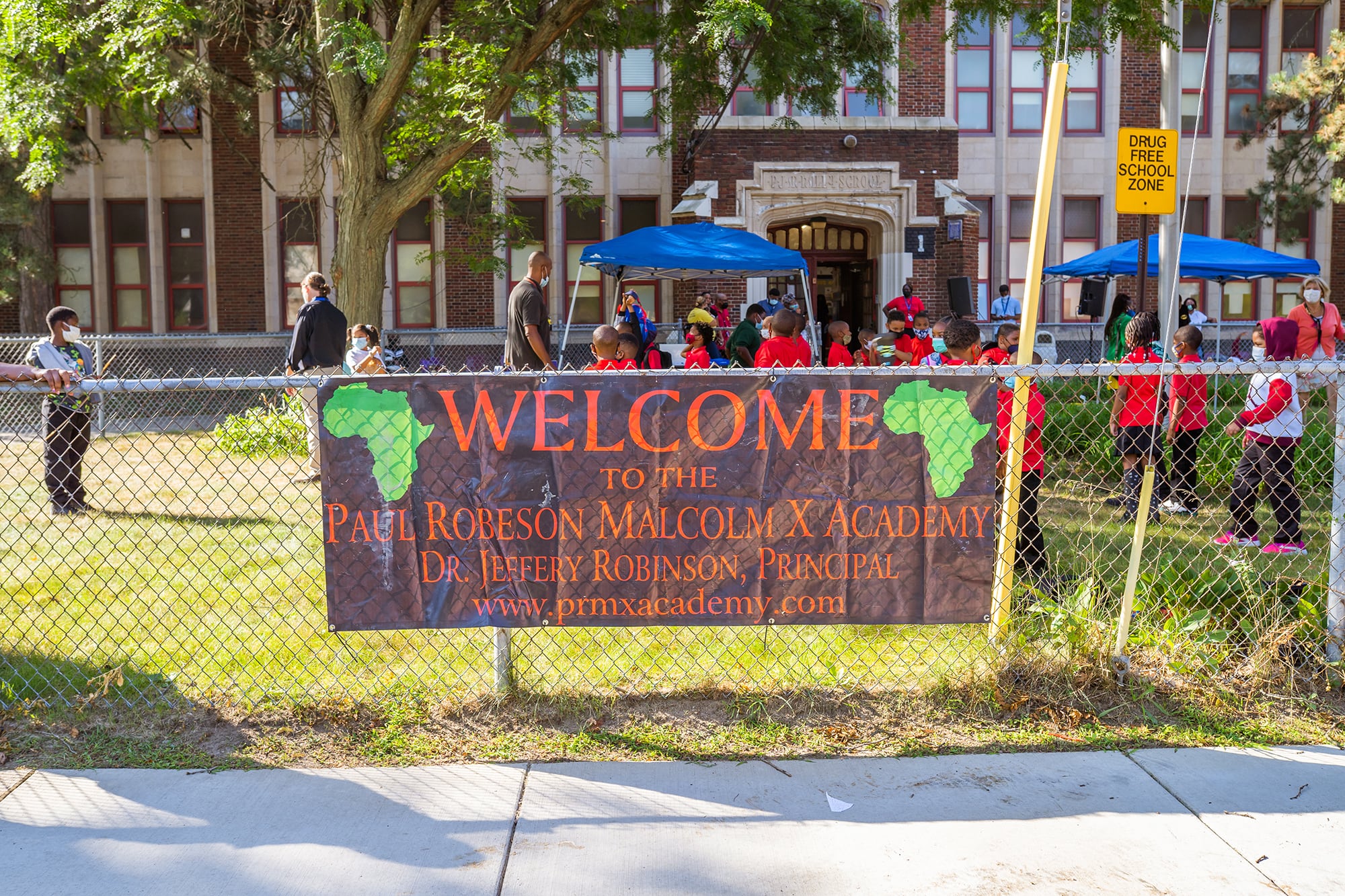 A large welcome sign hangs on the side of a fence in front of a school building and courtyard with young students outside.