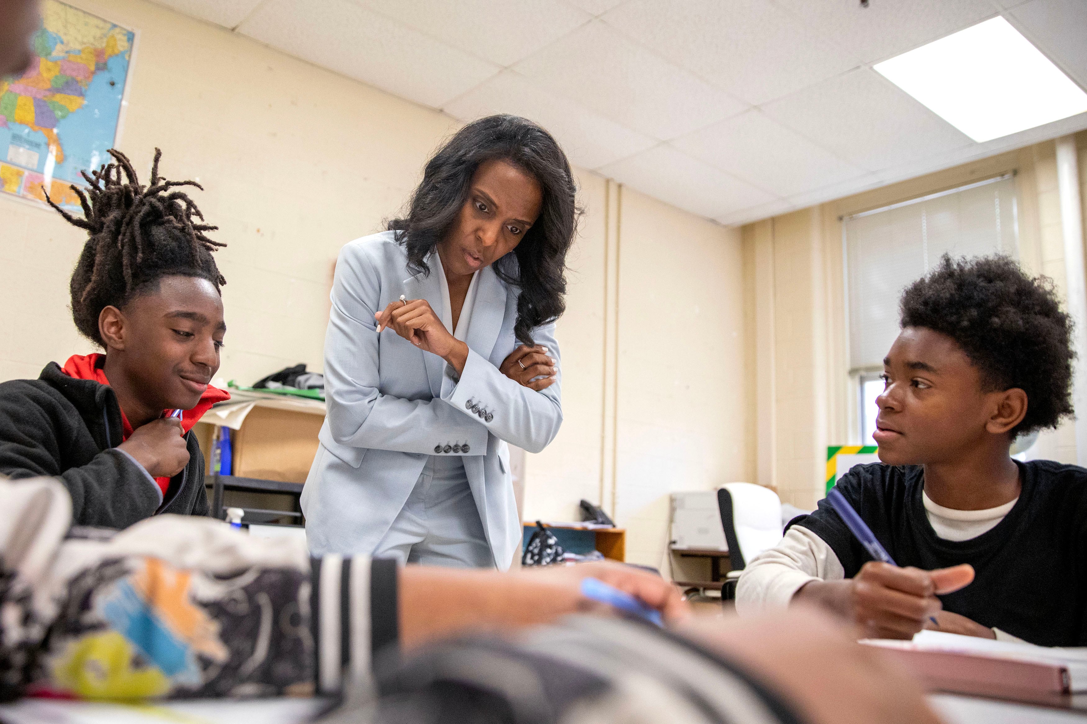 An adult wearing a light blue suit stands between two high middle school students who are sitting at their desks in a classroom.