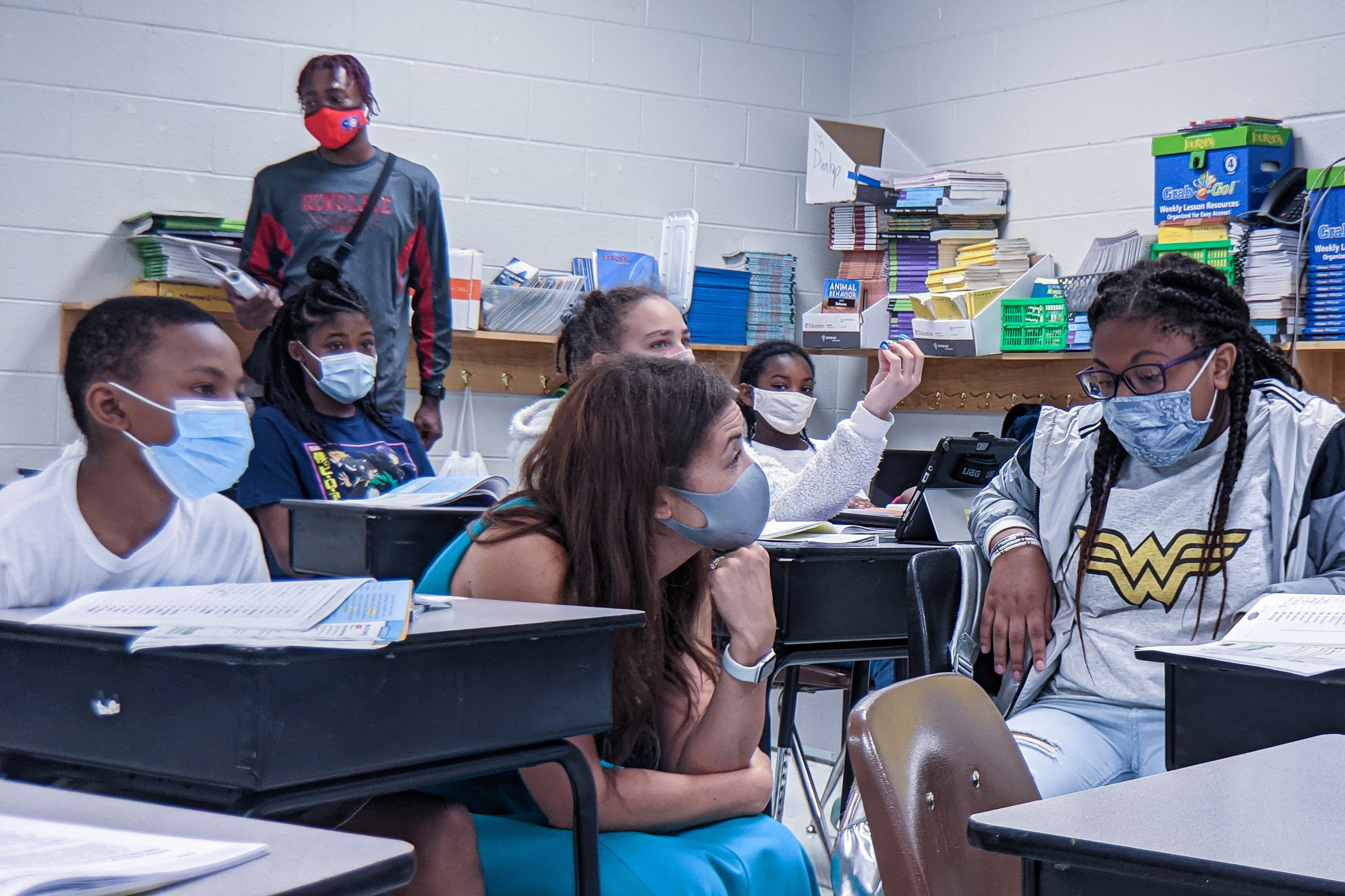 A woman crouches down to speak to a young girl in a classroom. They both are wearing masks.