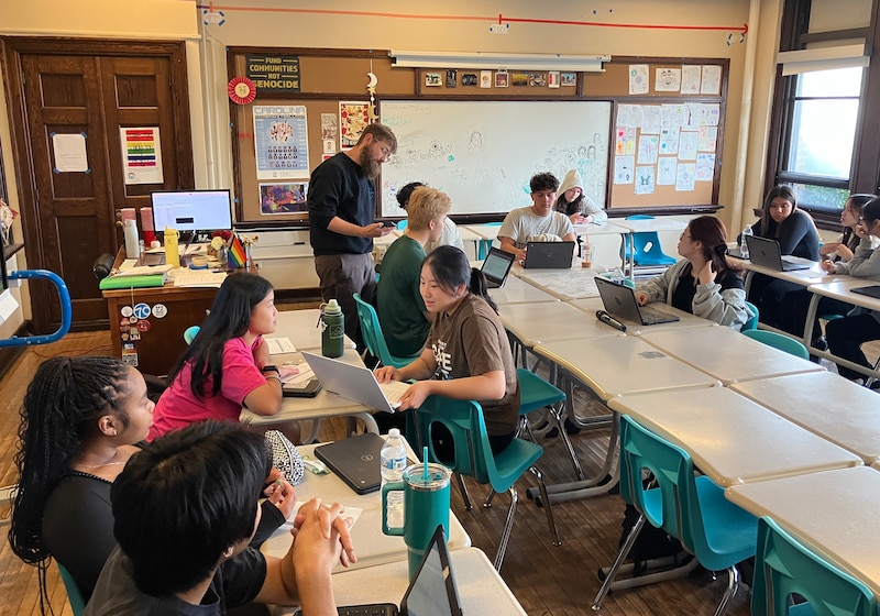 A teacher stands by a lot of students sitting in their chairs in a classroom.