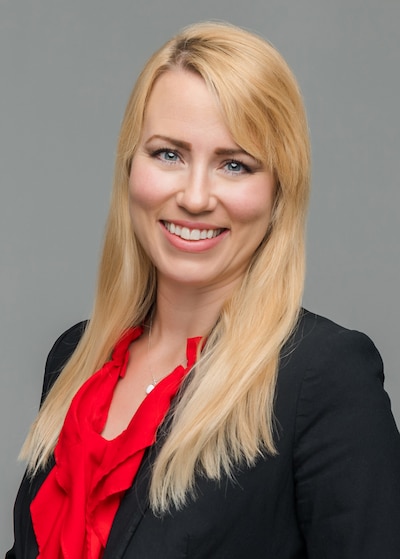 A woman with long blonde hair and wearing a red shirt and a dark suit jacket poses for a portrait in front of a grey wall.
