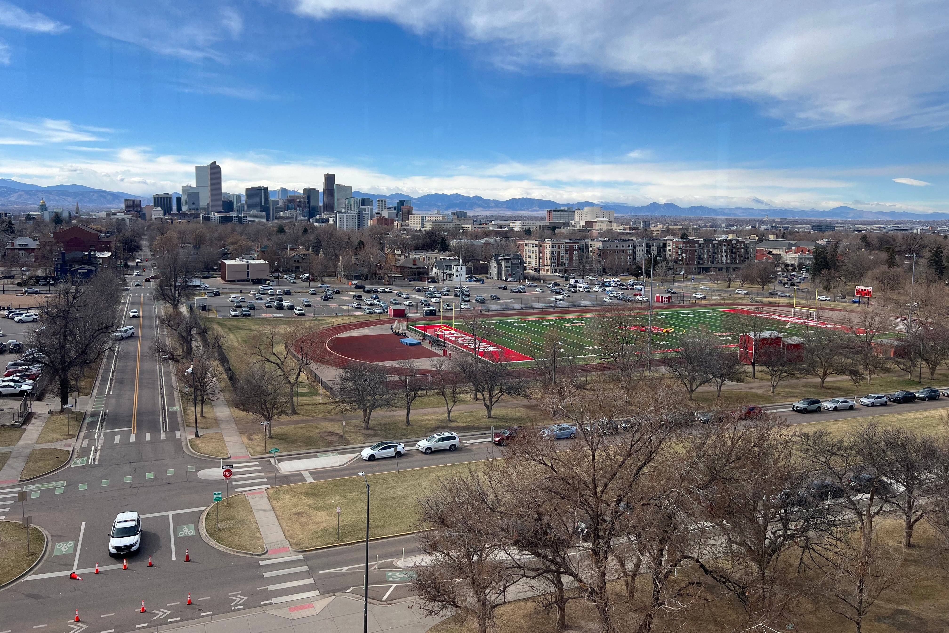 An aerial photograph of a high school track and football field in the middle of a couple of busy street with the skyline and mountains in the background.