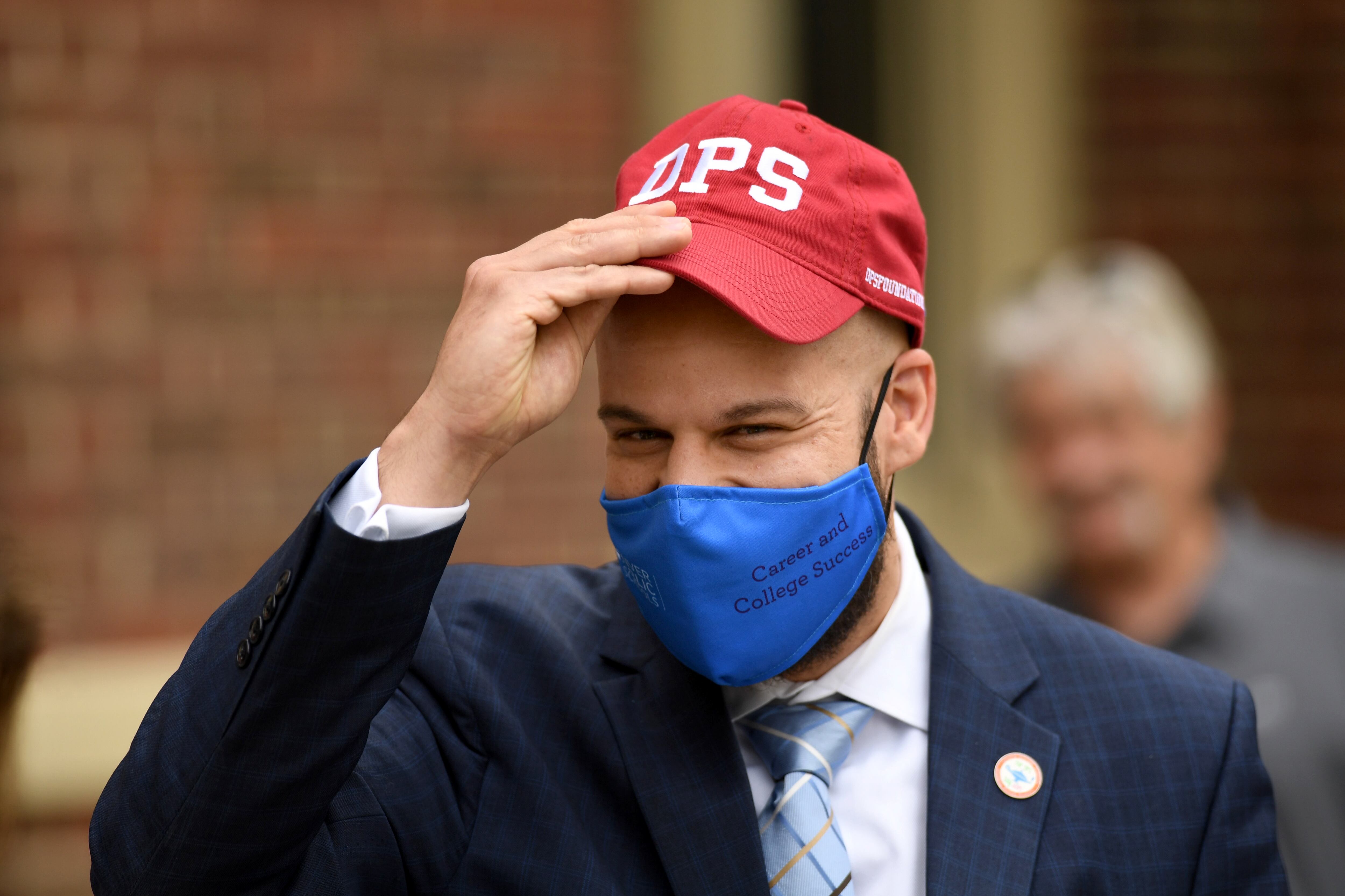 A close-up photo of the Denver Public Schools superintendent wearing a blue face mask. He is putting on a red baseball cap that says “DPS.”