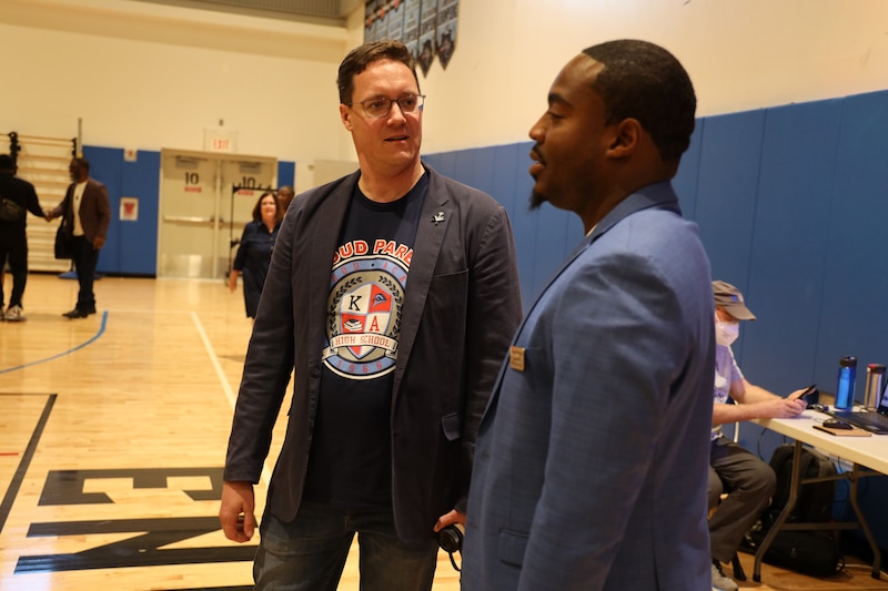 Two men wearing suit jackets speak at a high school gymnasium.