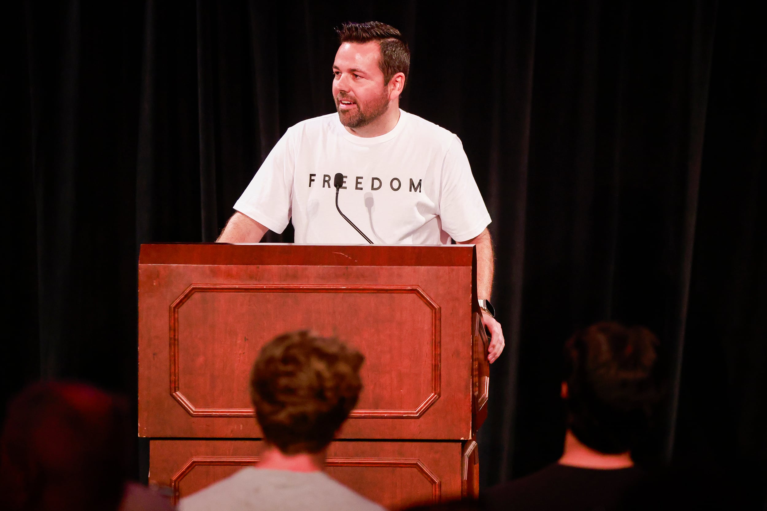 A photograph of a white man in a white tshirt speaks from behind a wooden podium with a few people in the foreground.