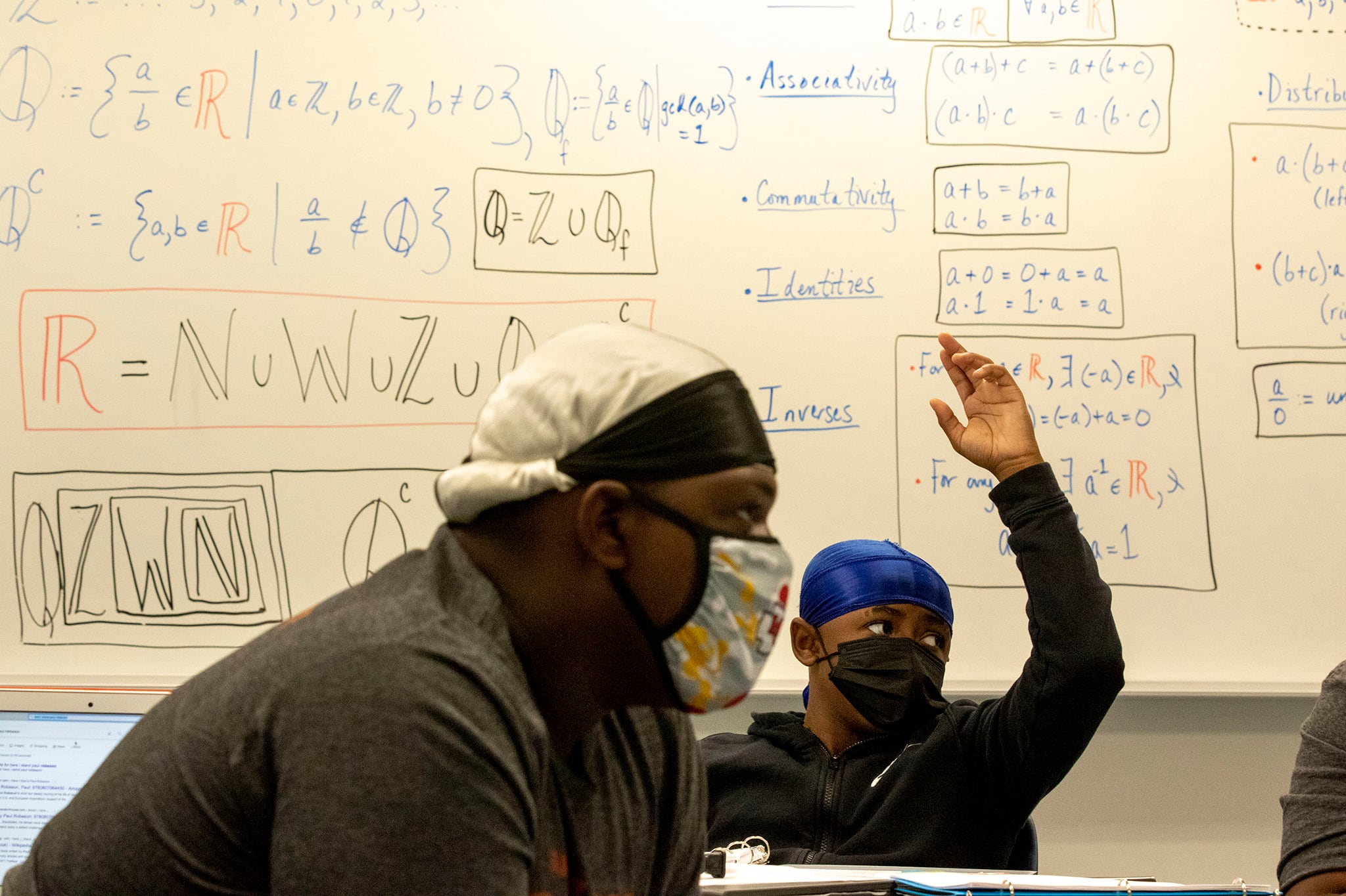 A Black student raises his hand in a classroom. A Black teacher can be seen in the foreground. Both are wearing face masks.