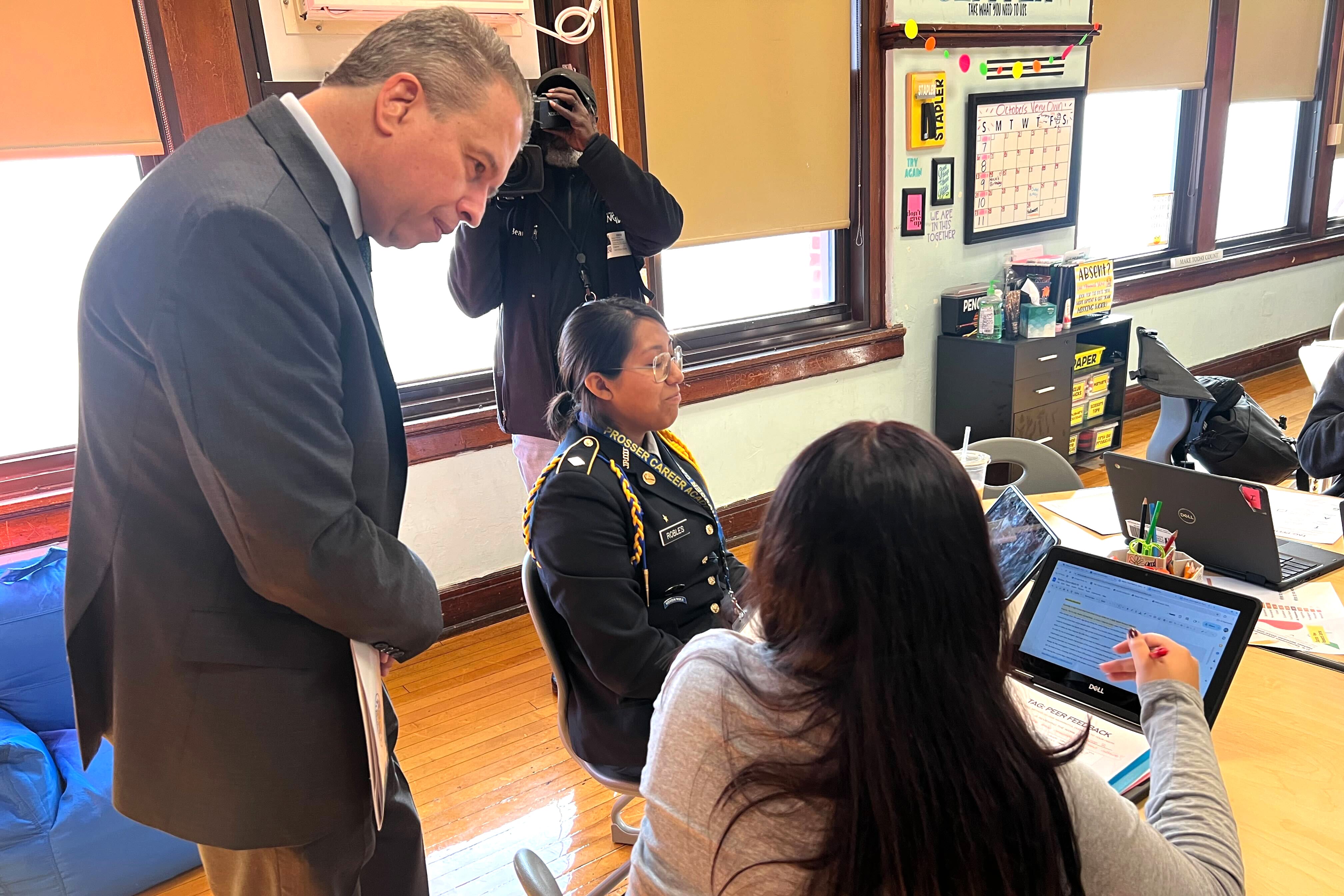 A man in a gray suit jacket leans over and talks to a female student with a laptop.