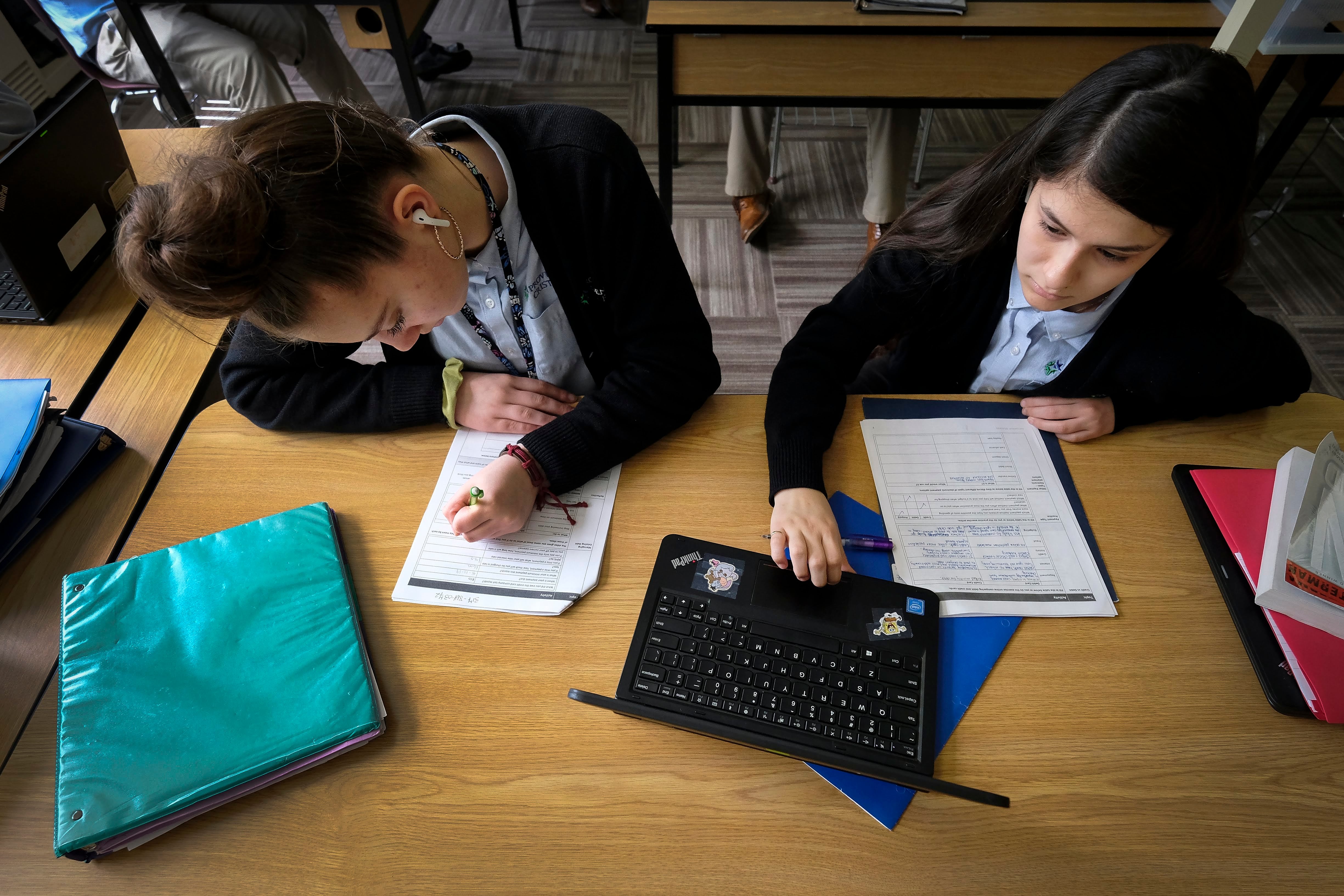 Two students sit at a desk working on assignments, while one uses the trackpad of a laptop in front of them.