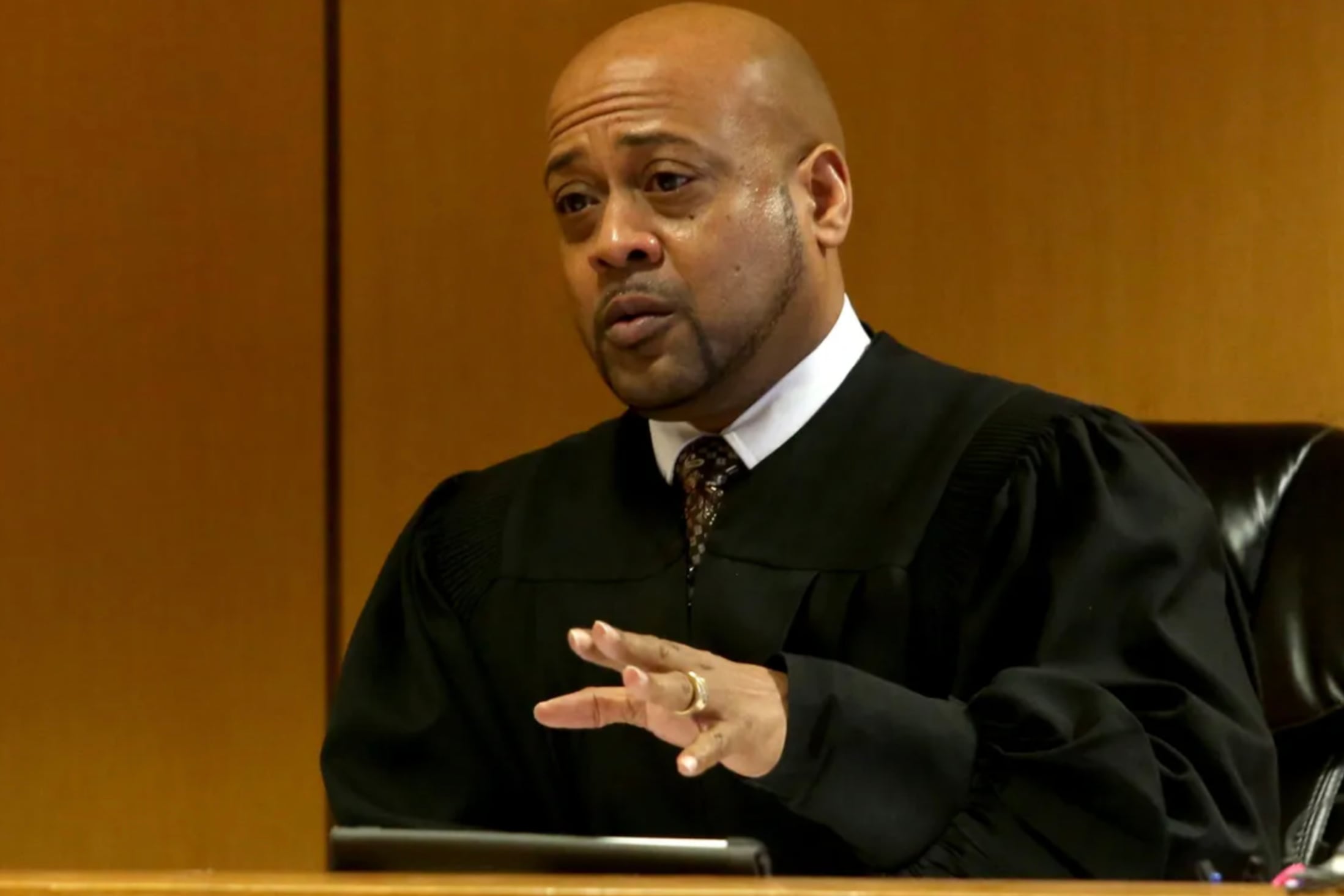 A man with no hair wearing a black judge robe sits at a chair in a courtroom with a wooden background.