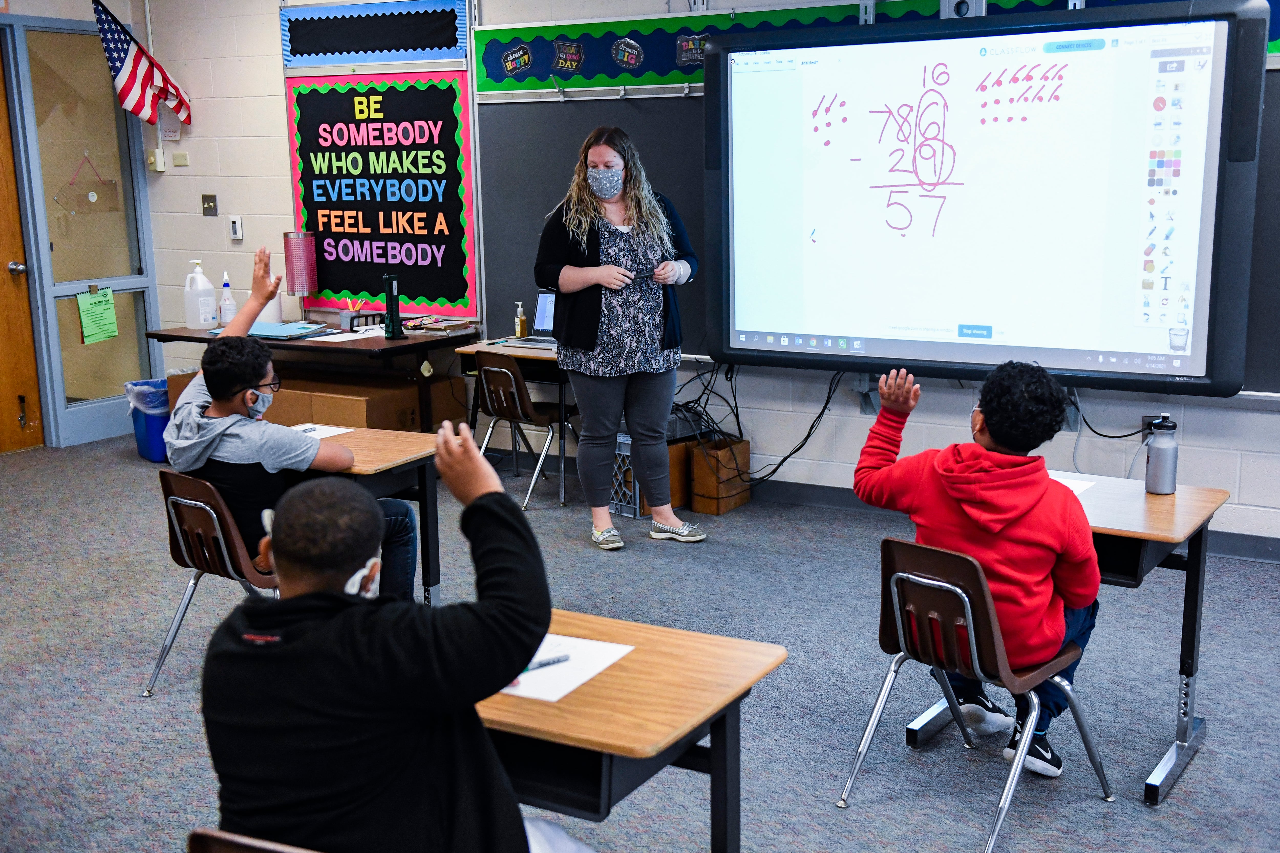 A teacher works through math problems on a smart board as her students raise their hands at their desks.