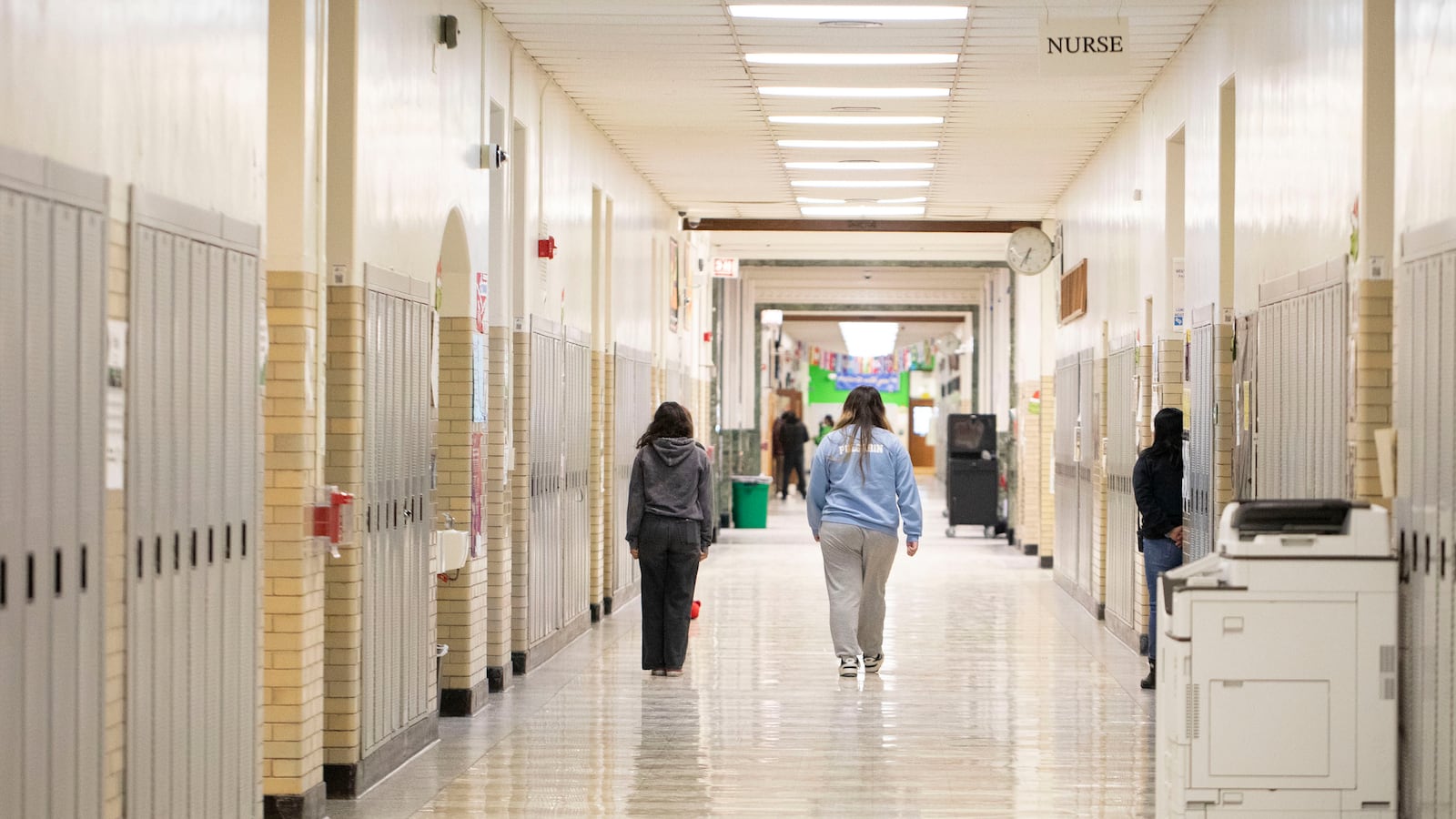 A photograph of high school students walking down a hallway with lockers on each side.