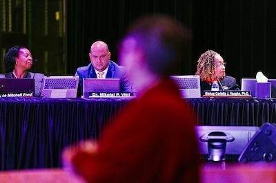 Several people are sitting behind a table during a Detroit school board meeting as a woman wearing a red shirt passes by.