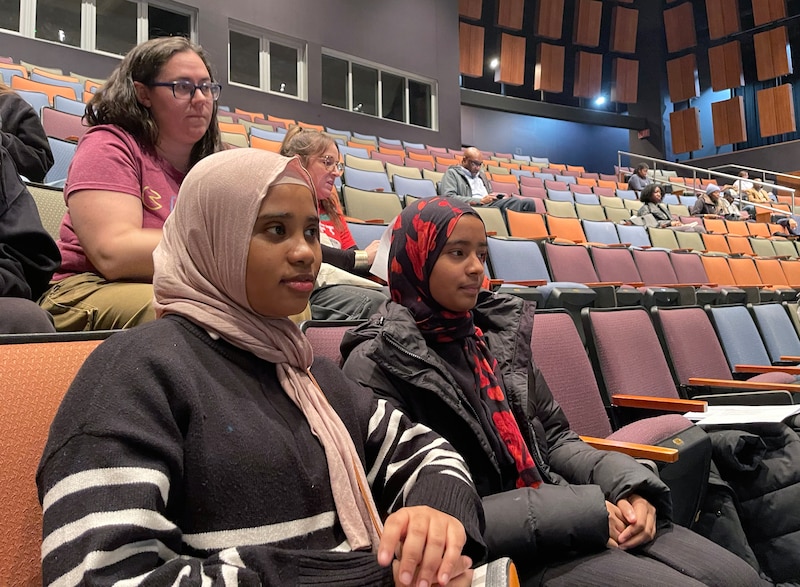 A photograph of two recently graduated high school students both wearing head scarves, sit next to each other in a large auditorium. There are a few people sitting behind them.