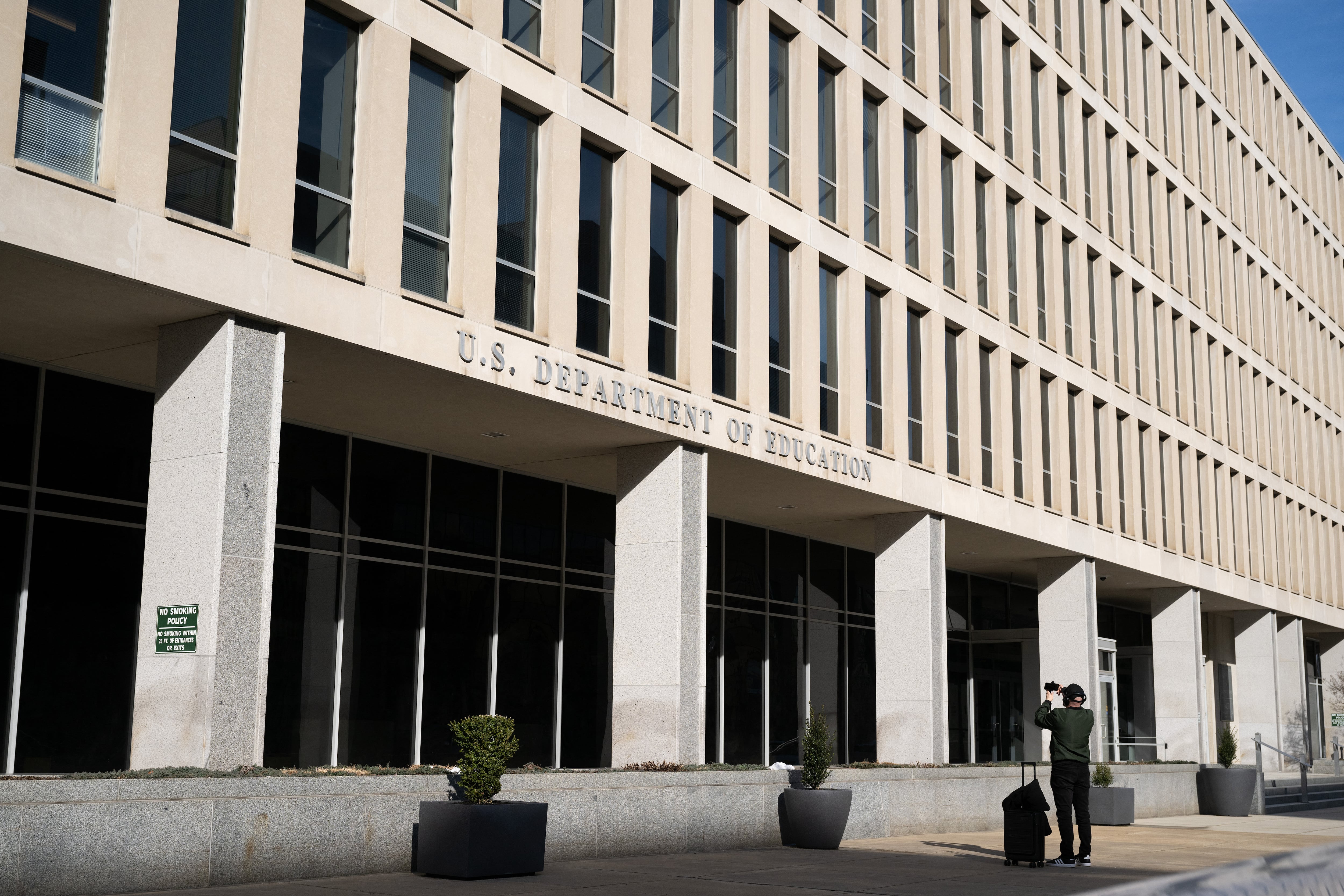 A person stands in front of a large stone building with a sign that reads "U.S. Department of Education."