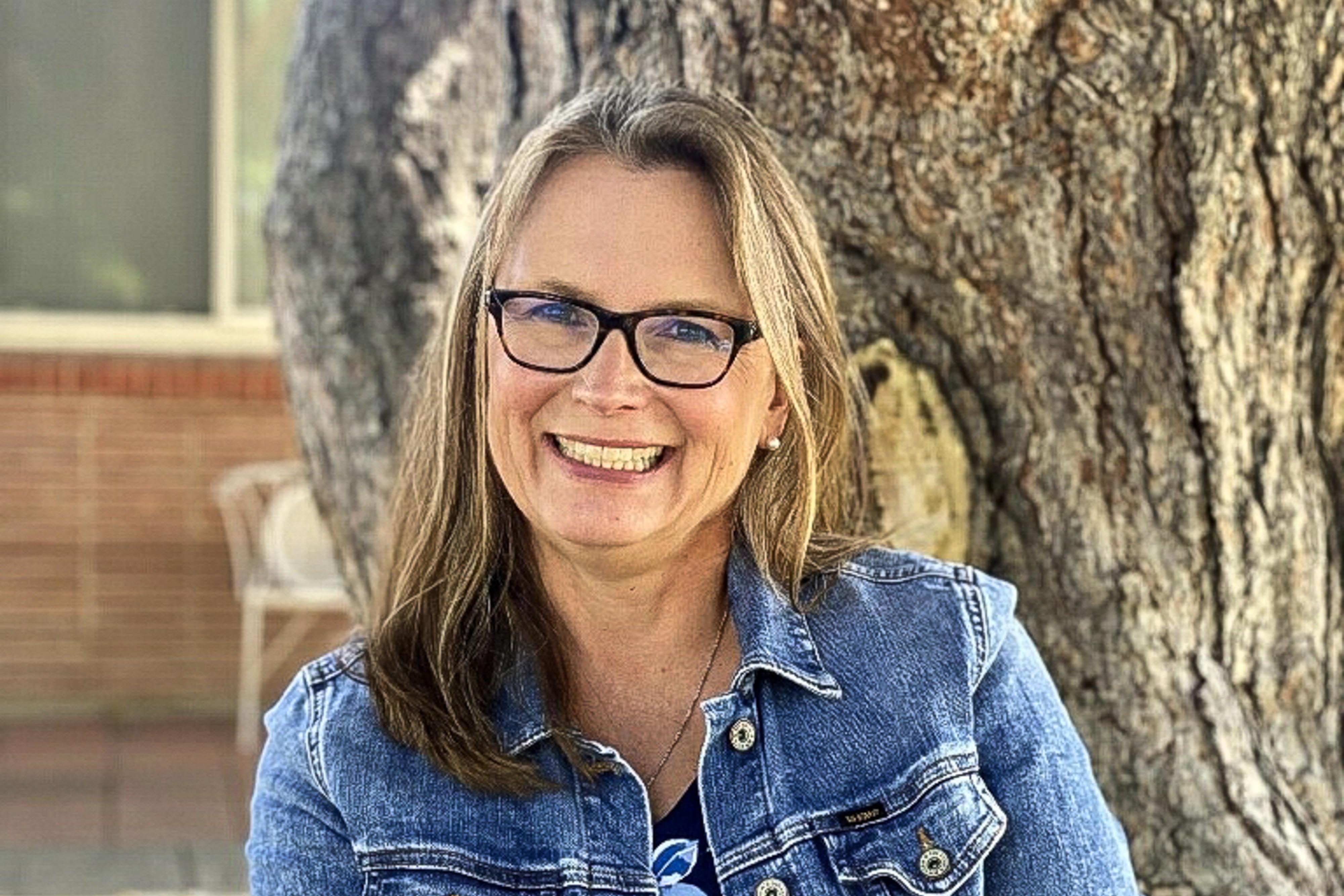 A portrait of Denver school board candidate Carrie Olson, who is sitting against a tree wearing denim jacket and glasses.