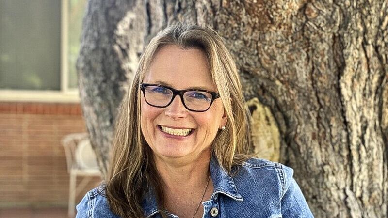 A portrait of Denver school board candidate Carrie Olson, who is sitting against a tree wearing denim jacket and glasses.