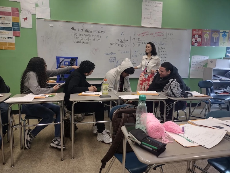 Four students sit at desk while another in a white top stands behind them.