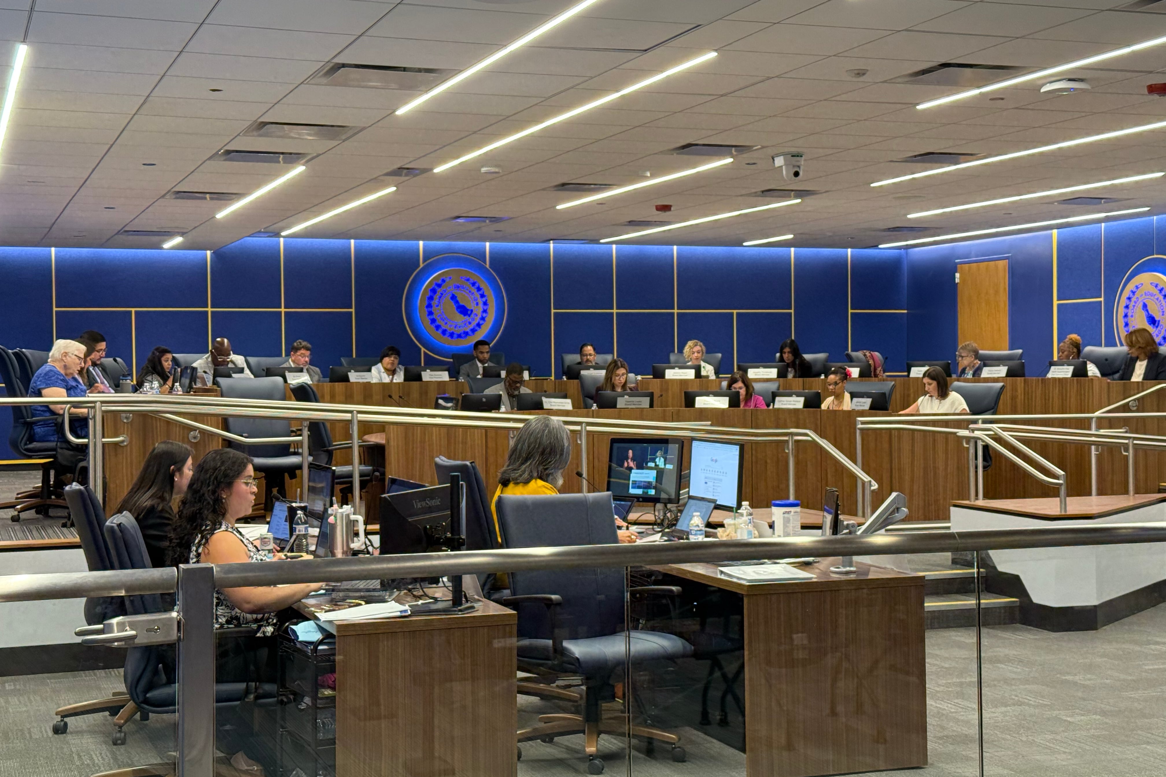 A photograph of a large school board meeting in a conference room.